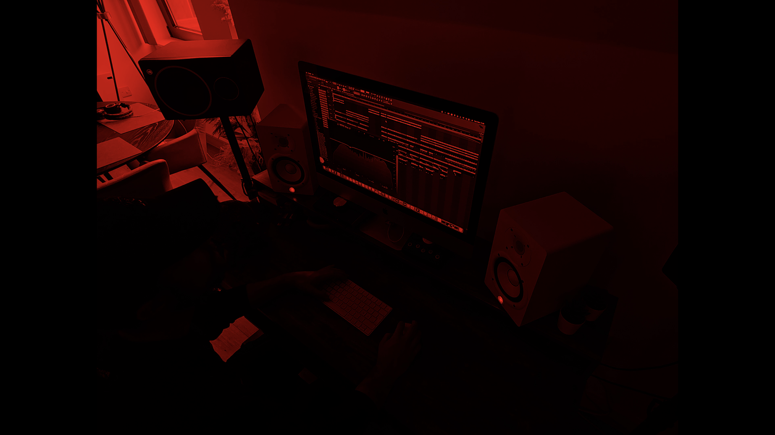 A dark room lit by red light showing a computer monitor displaying audio editing software, surrounded by studio speakers and a person working at a desk with a keyboard and mouse.