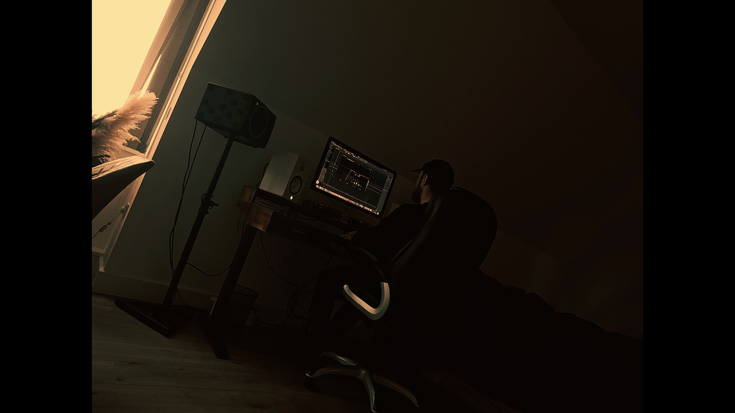 Person working on a computer in a dimly lit room, illuminated by a window light, with audio equipment nearby.