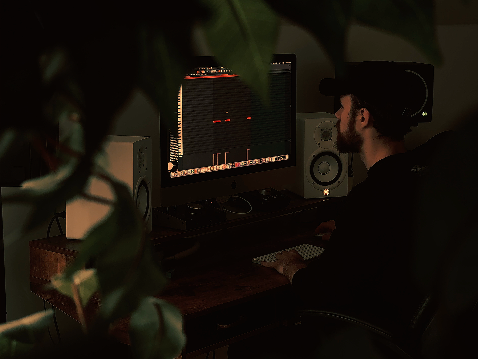 A man working on a music production computer setup with speakers, stylus, and mixing software visible on the screen, partially obscured by blurred leaves in the foreground.