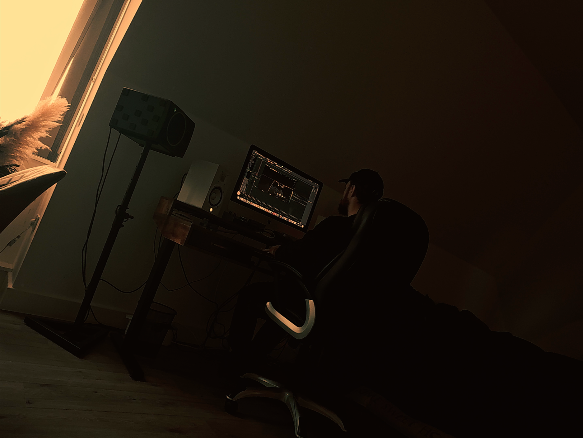 A person working on a computer with two speakers in a dimly lit room near a window.