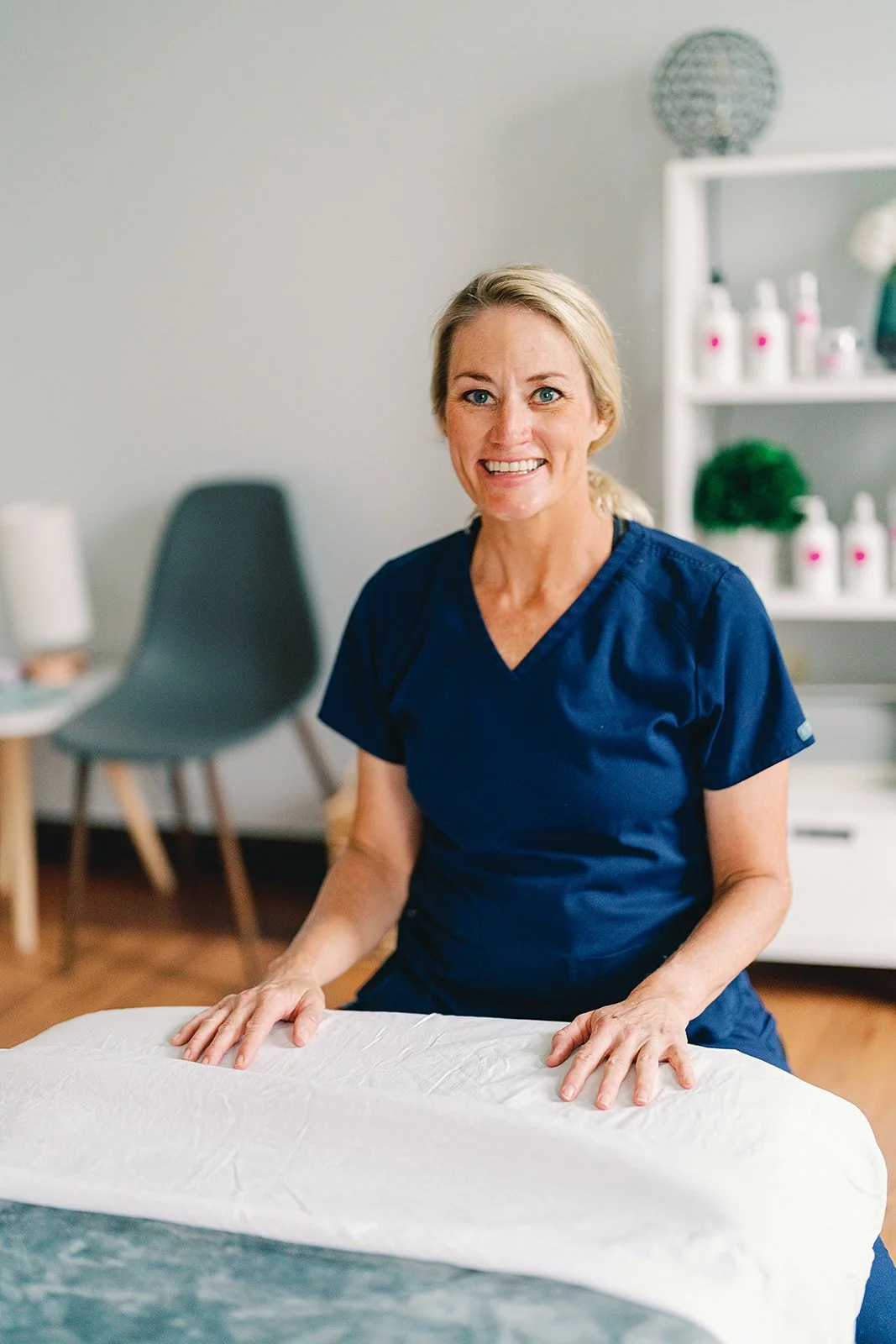 A woman in a navy blue uniform smiling as she presses her hands on a massage table in a wellness or massage therapy room.