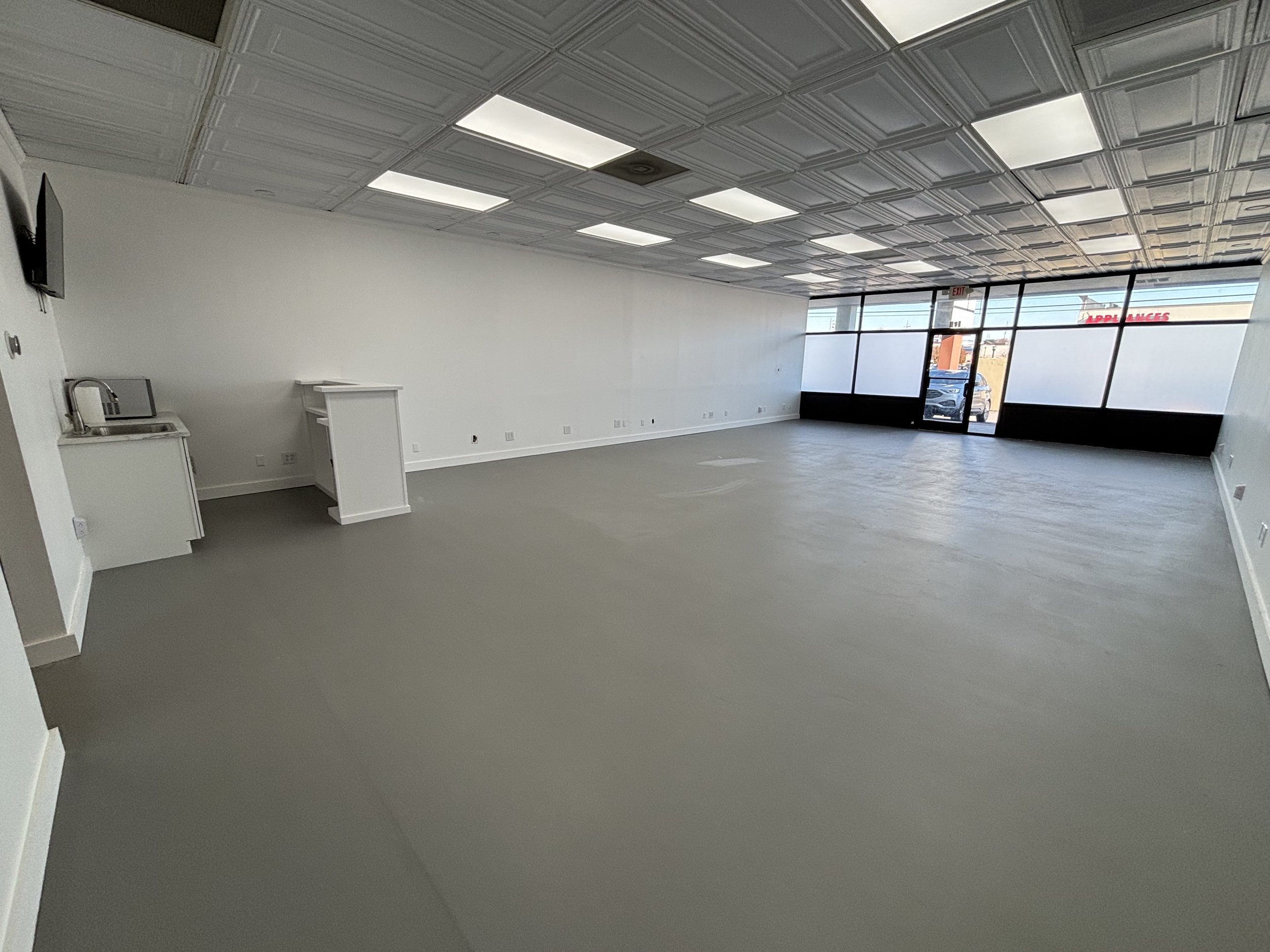 Empty commercial space with large front windows, white walls, a gray floor, and ceiling tiles. A small kitchenette area with a microwave, sink, and counter on the left side.
