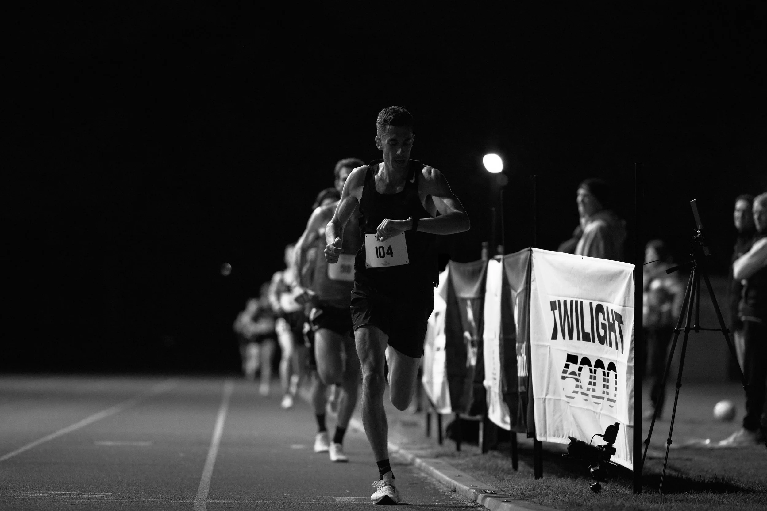 Nighttime marathon race with runners, including a man wearing bib number 104, running on a track near a Twilight 500 banner, with crowd and race officials in the background.