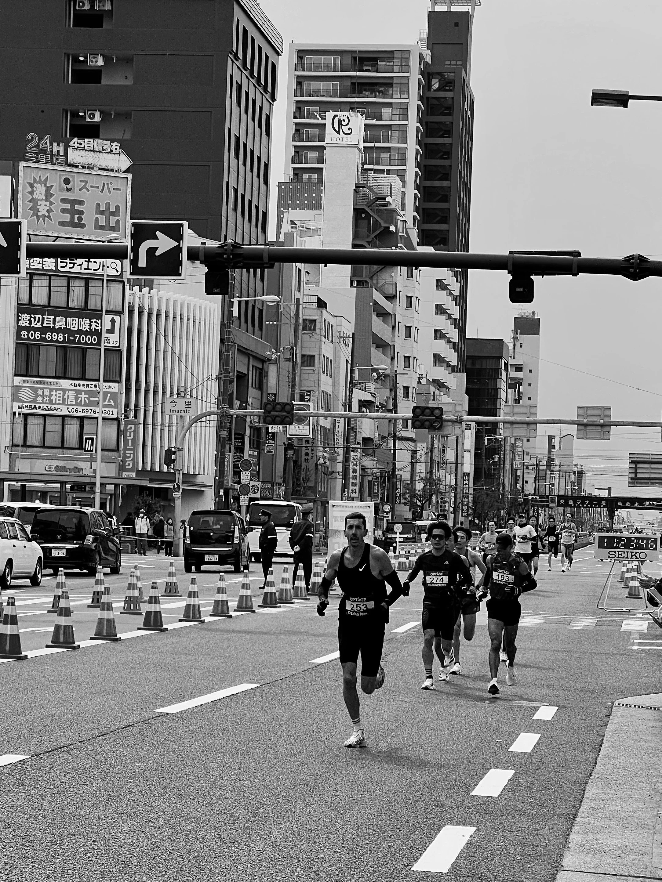 A marathon race with multiple runners on a city street, surrounded by tall buildings and traffic cones, with a cityscape in the background.