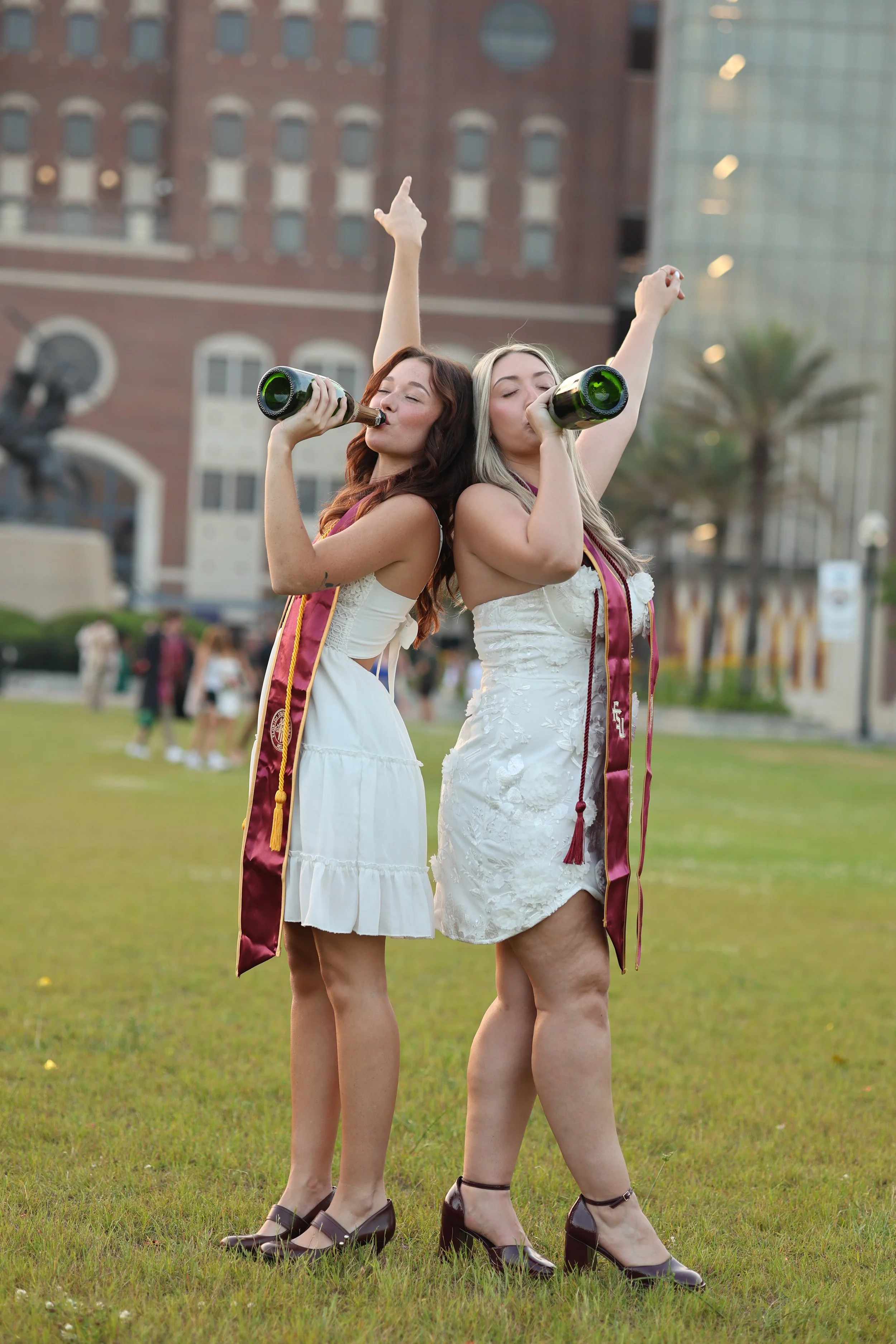 Two women at a graduation celebration, wearing white dresses and maroon graduation stoles, standing on a grass field and drinking from bottles, with their eyes closed and arms raised, in front of a modern building with large glass windows and a statu