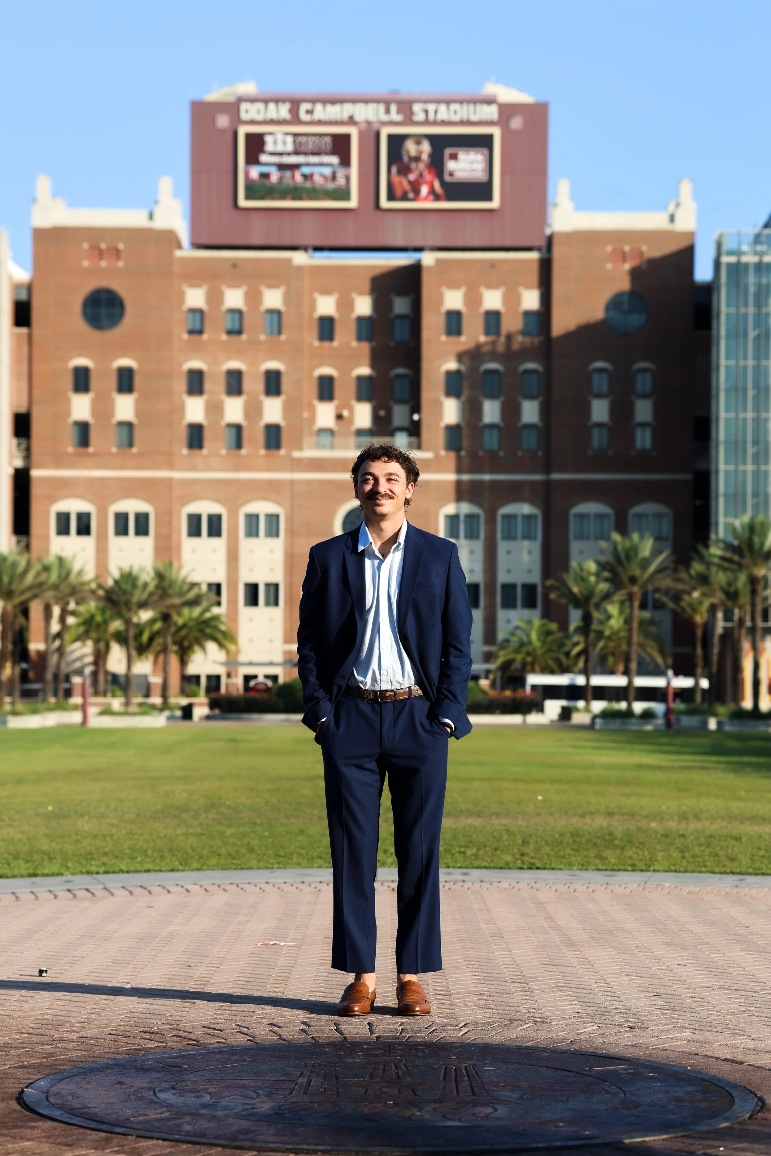 A man in a navy suit standing on a circular brick platform outdoors, with a green lawn and palm trees in the background, and a brick building with a large digital scoreboard behind him.