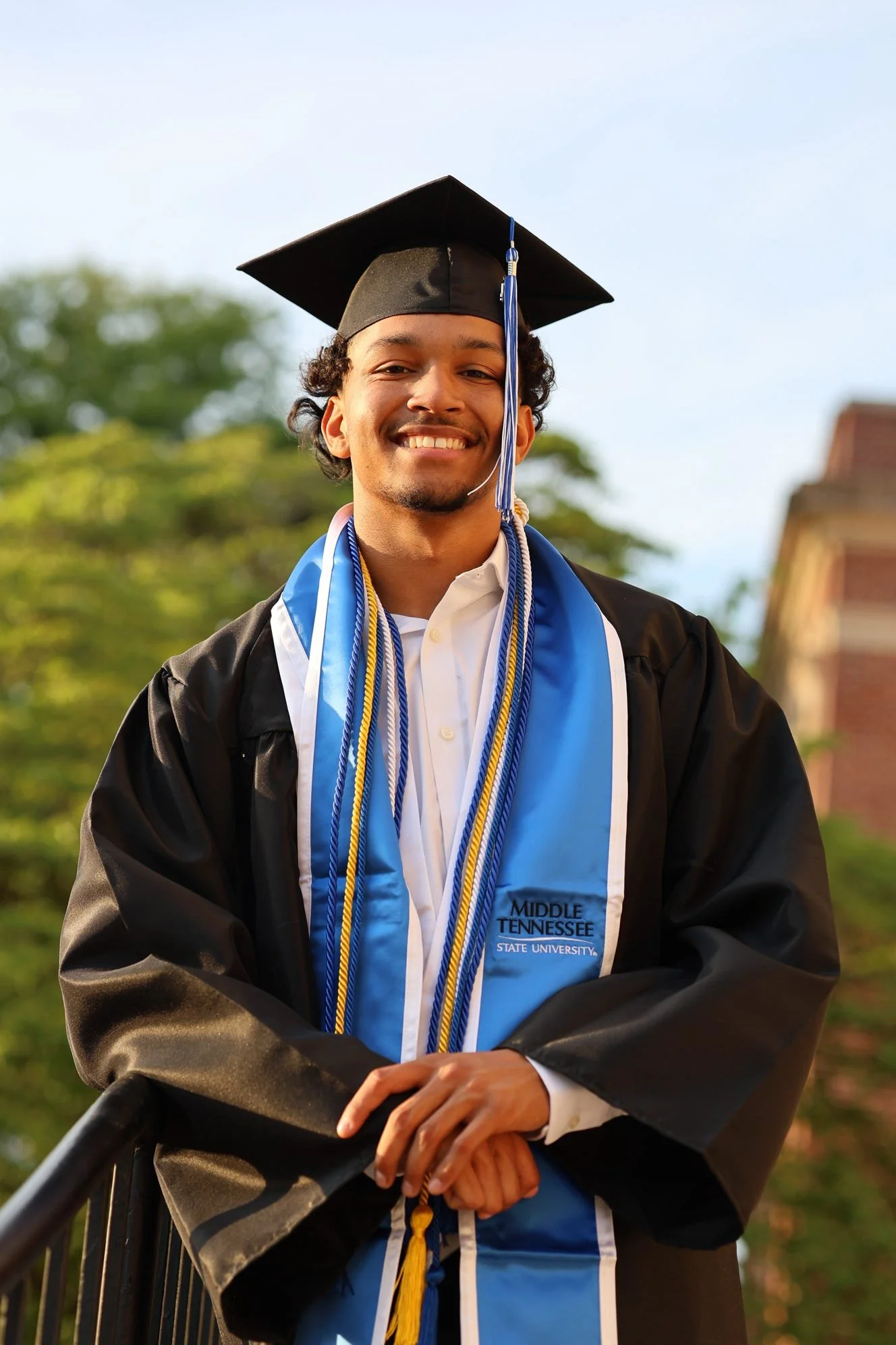 Graduate in black cap and gown with blue and white stole, smiling outdoors.