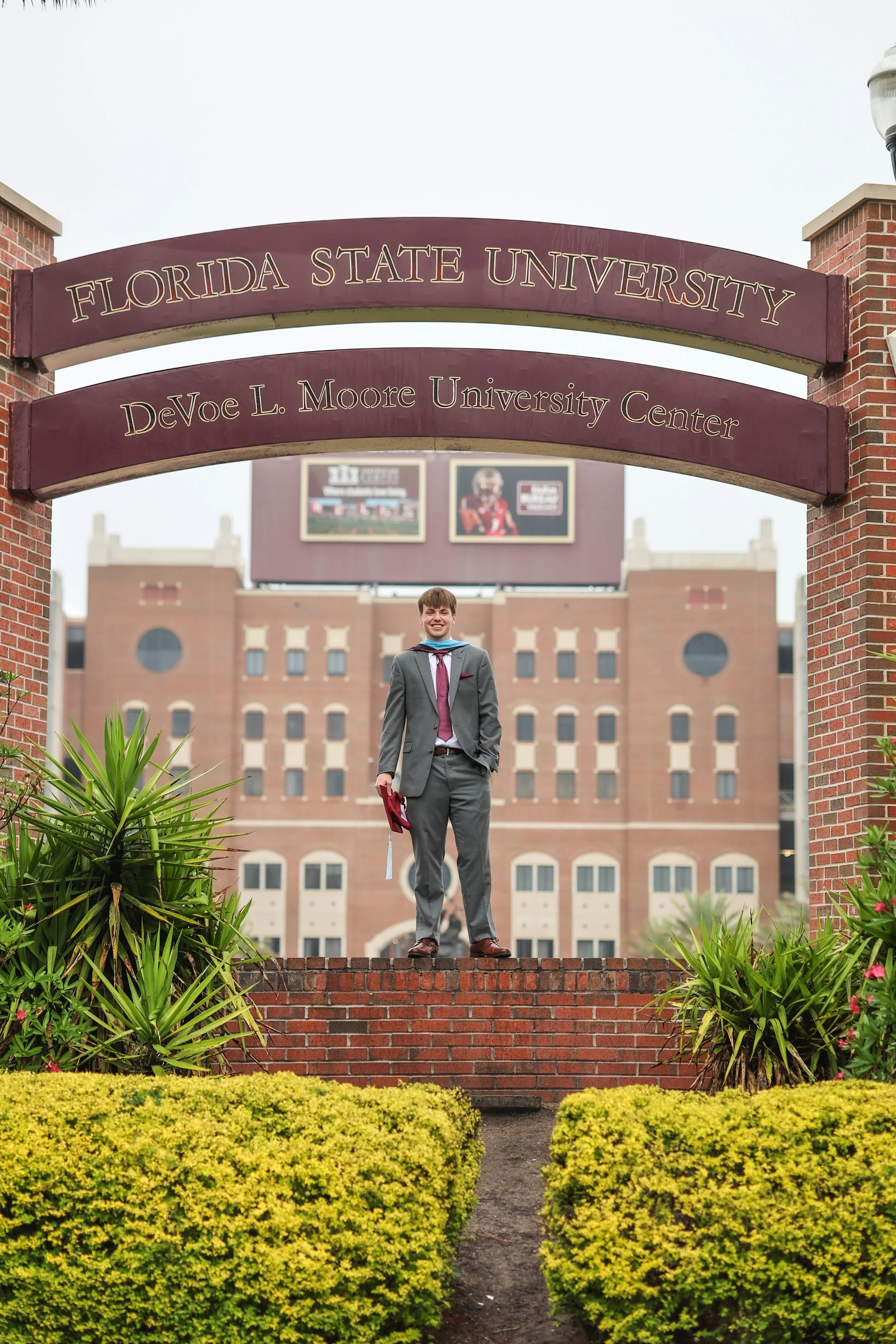 A young man in a graduation cap and gown standing on a brick ledge in front of Florida State University, with the DeVoe L. Moore University Center building in the background.
