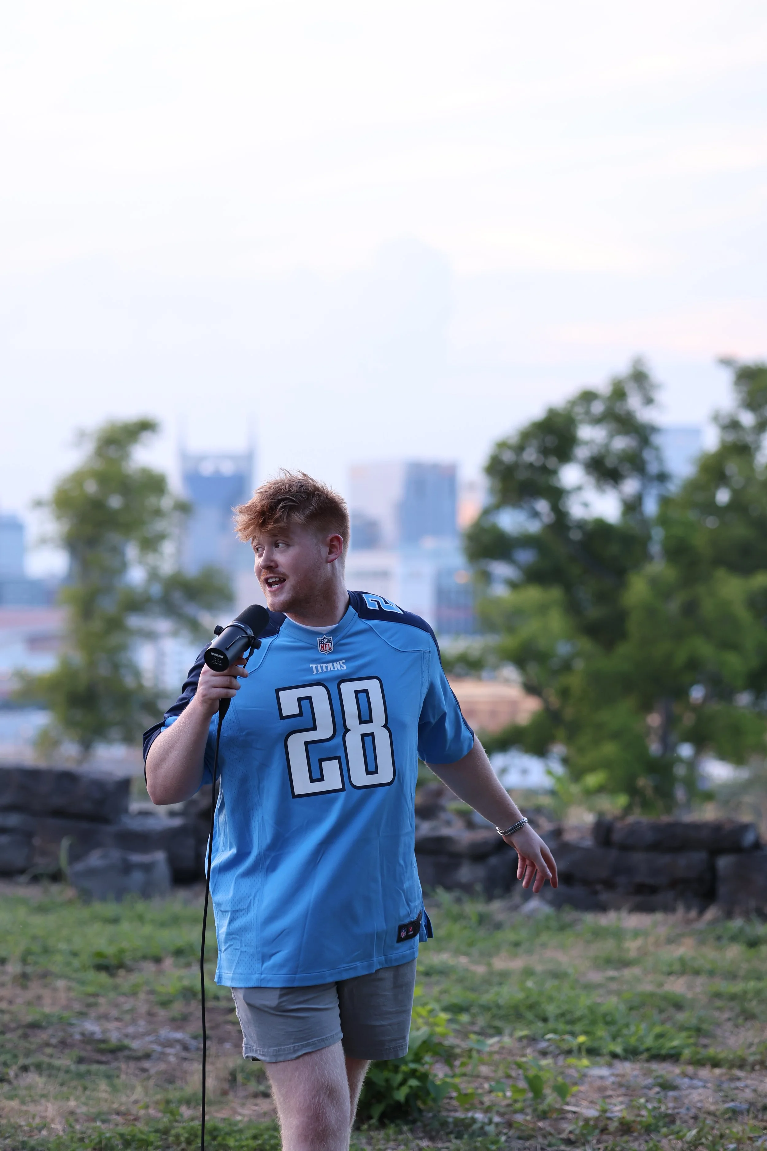 A young man wearing a blue Tennessee Titans football jersey with the number 28, holding a microphone and speaking outdoors.