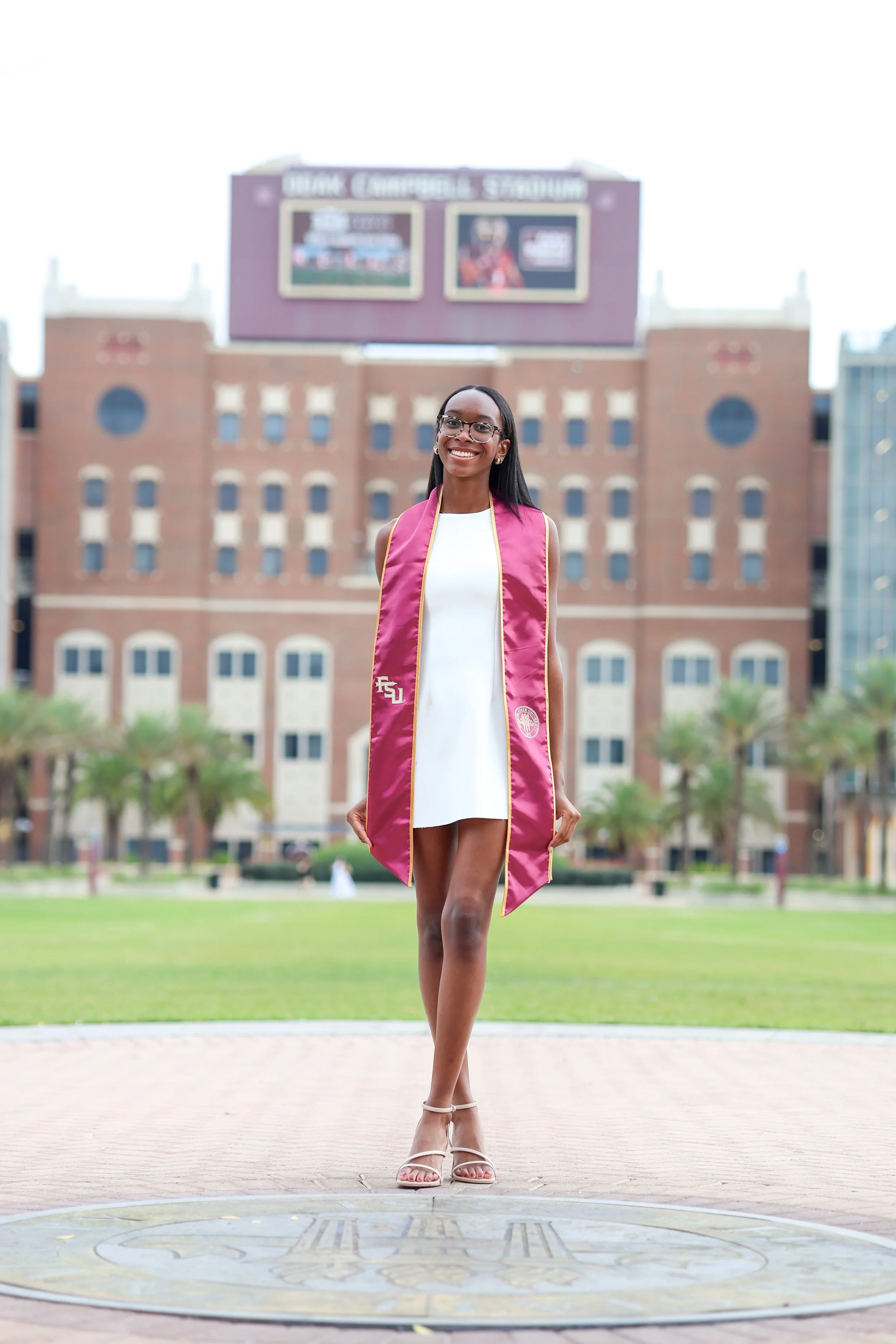A woman in a white dress and pink graduation gown standing outdoors in front of a university building and smiling.
