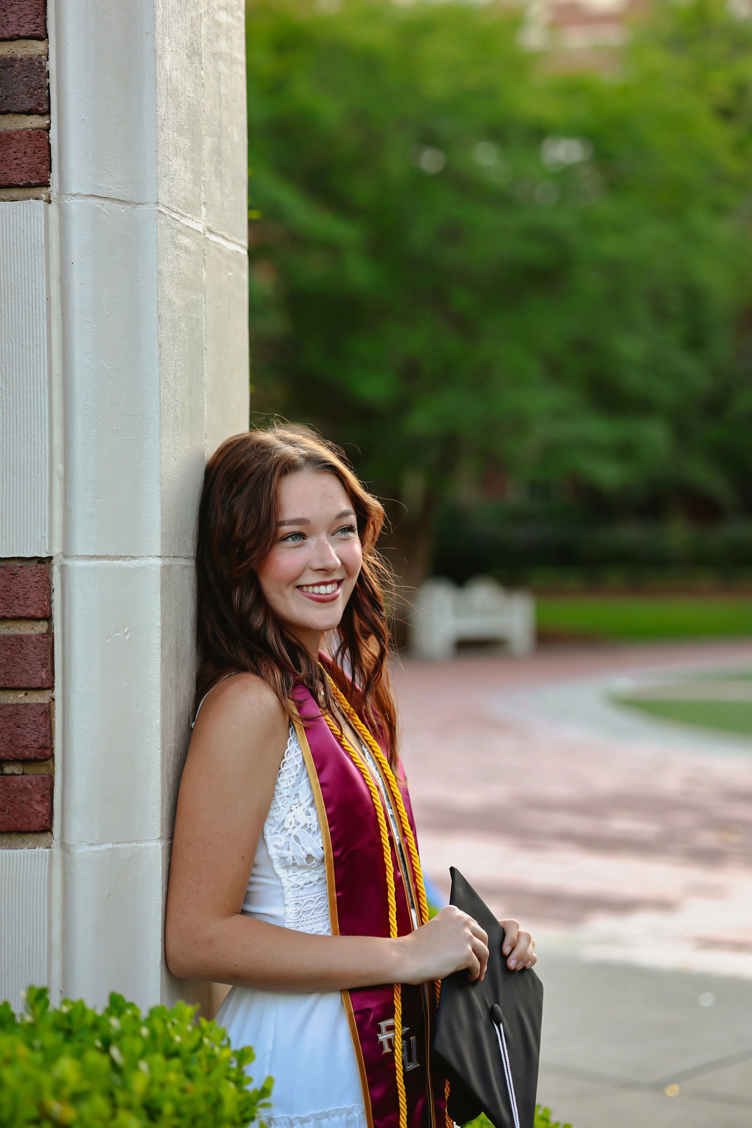 Young woman smiling, wearing a white dress and maroon graduation sash, holding a graduation cap and standing outdoors next to a building with trees in the background.