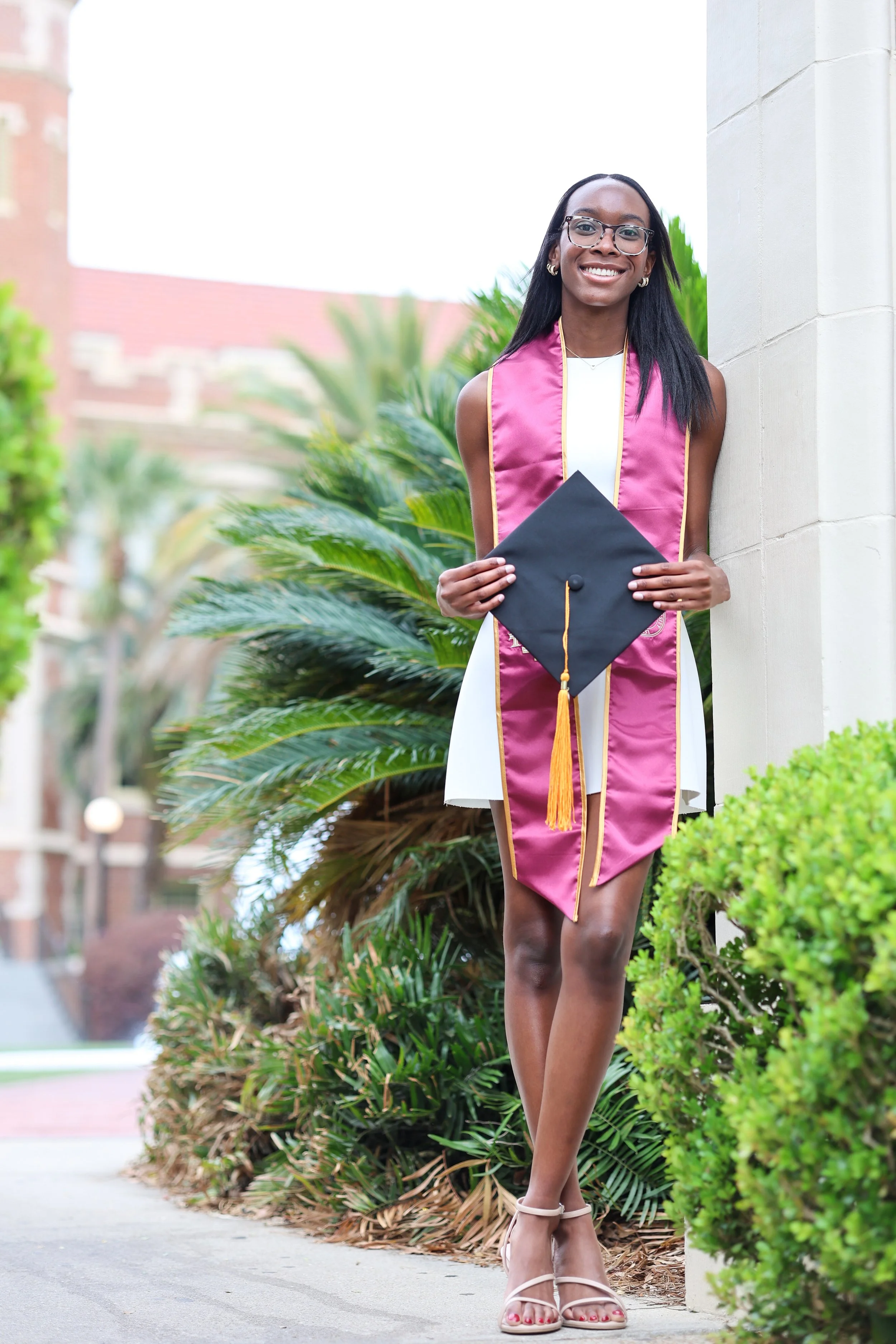 A young woman in a white dress and pink graduation stole with gold trim stands outdoors beside a stone wall, holding a black graduation cap with a gold tassel, smiling at the camera, with lush greenery and a brick building in the background.