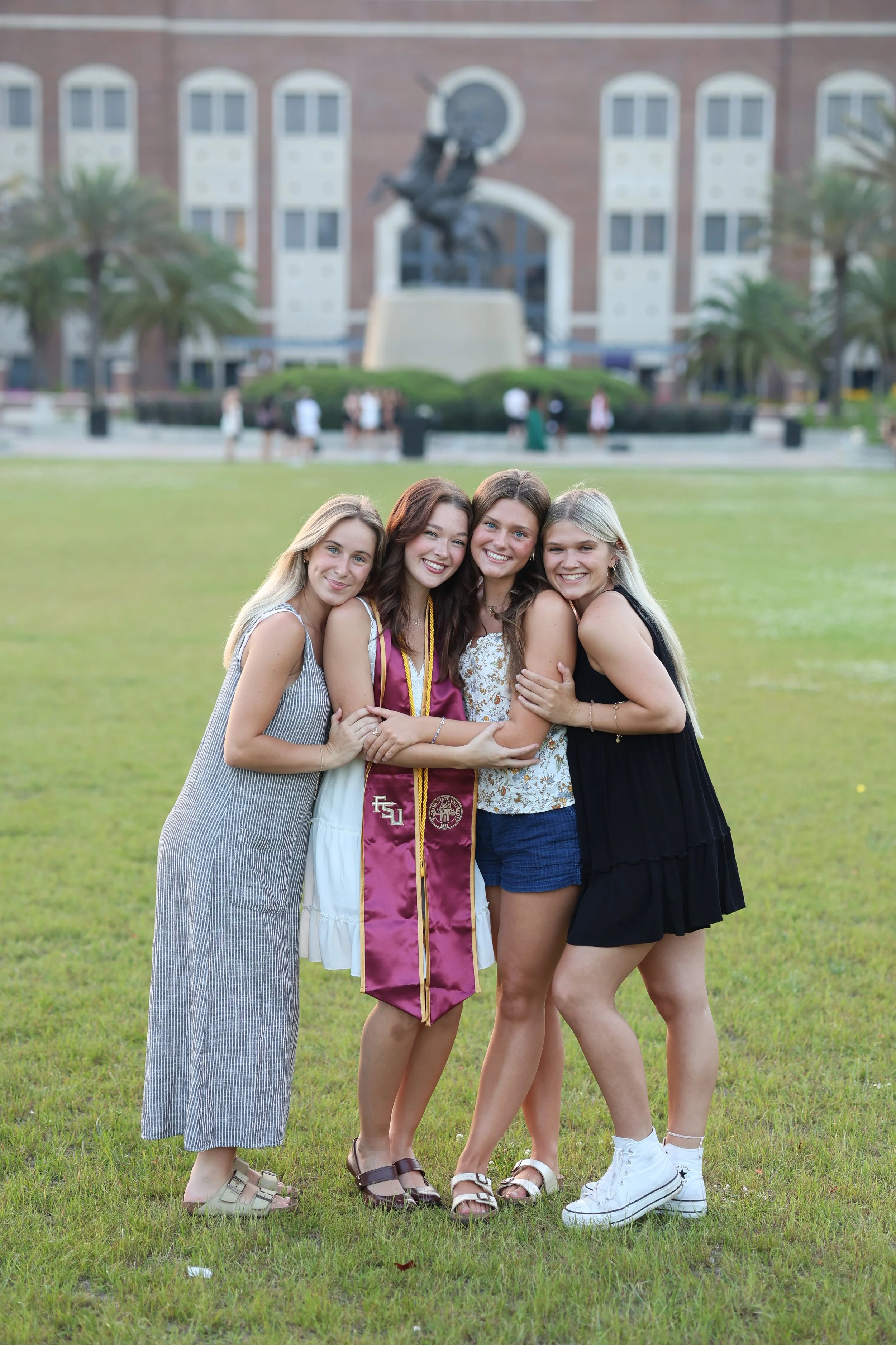 Four young women standing on grass, with one in a graduation gown, smiling and hugging each other, with a building and a statue in the background.