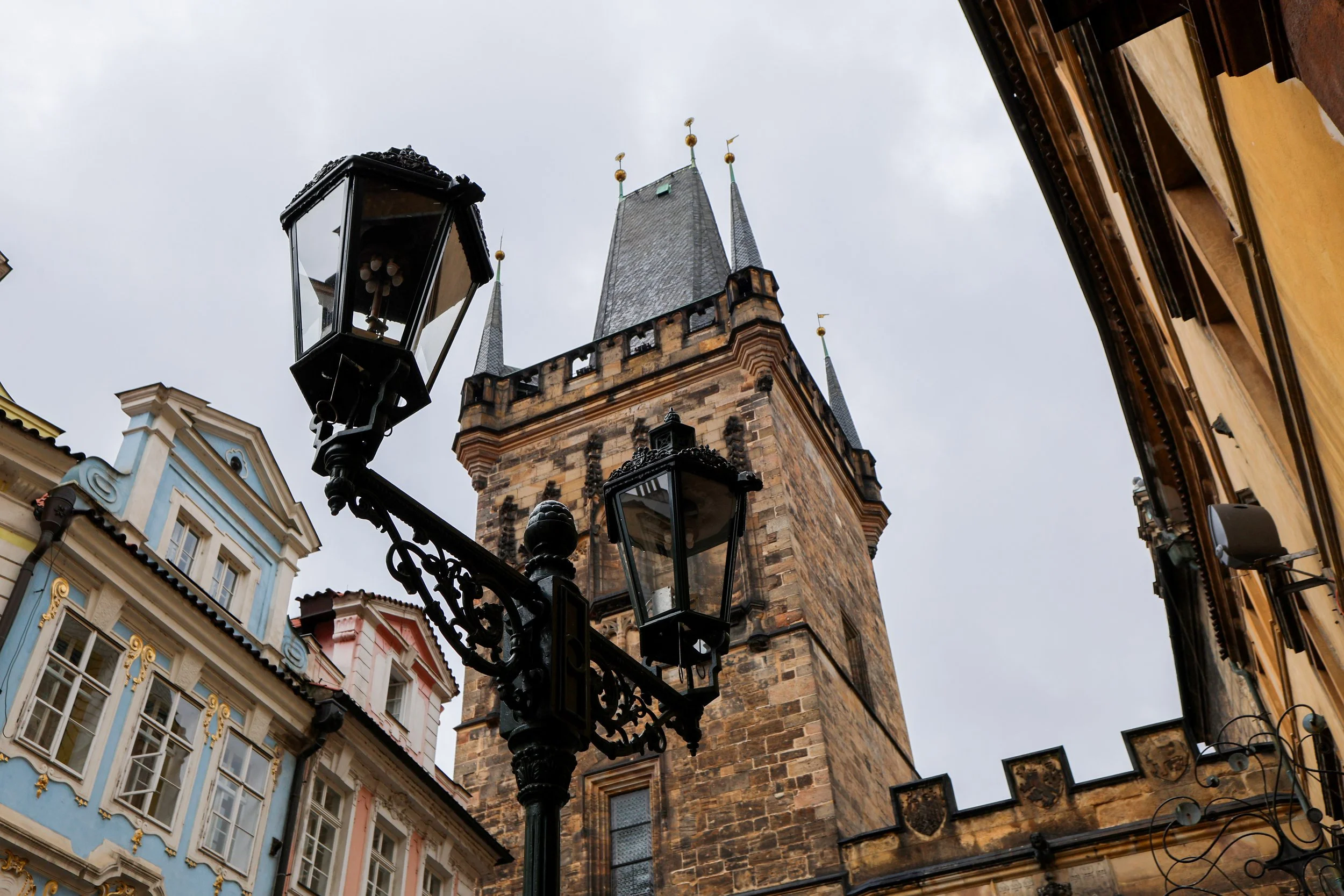 Historic stone tower with pointed spires and gold accents, flanked by ornate vintage street lamps, and colorful European buildings with detailed facades and windows.