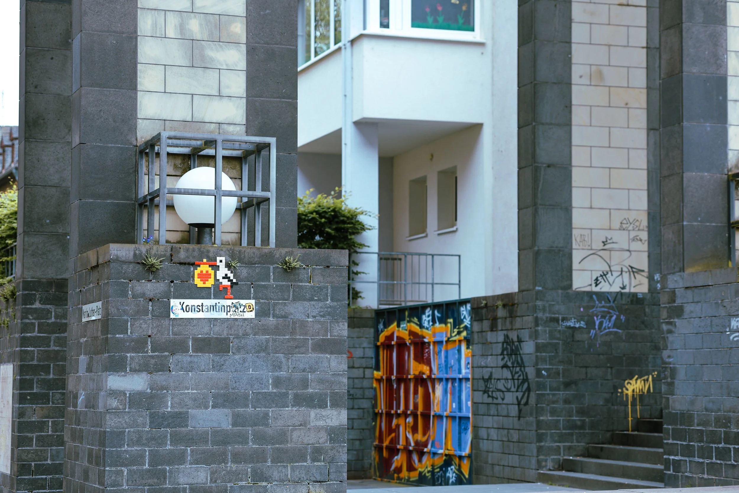 Urban scene featuring a brick pillar with a modern white spherical light fixture enclosed in a metal cage, graffiti-covered walls, and a mural painted on a closed gate with orange, blue, and yellow colors. There are also stairs and a building with wh