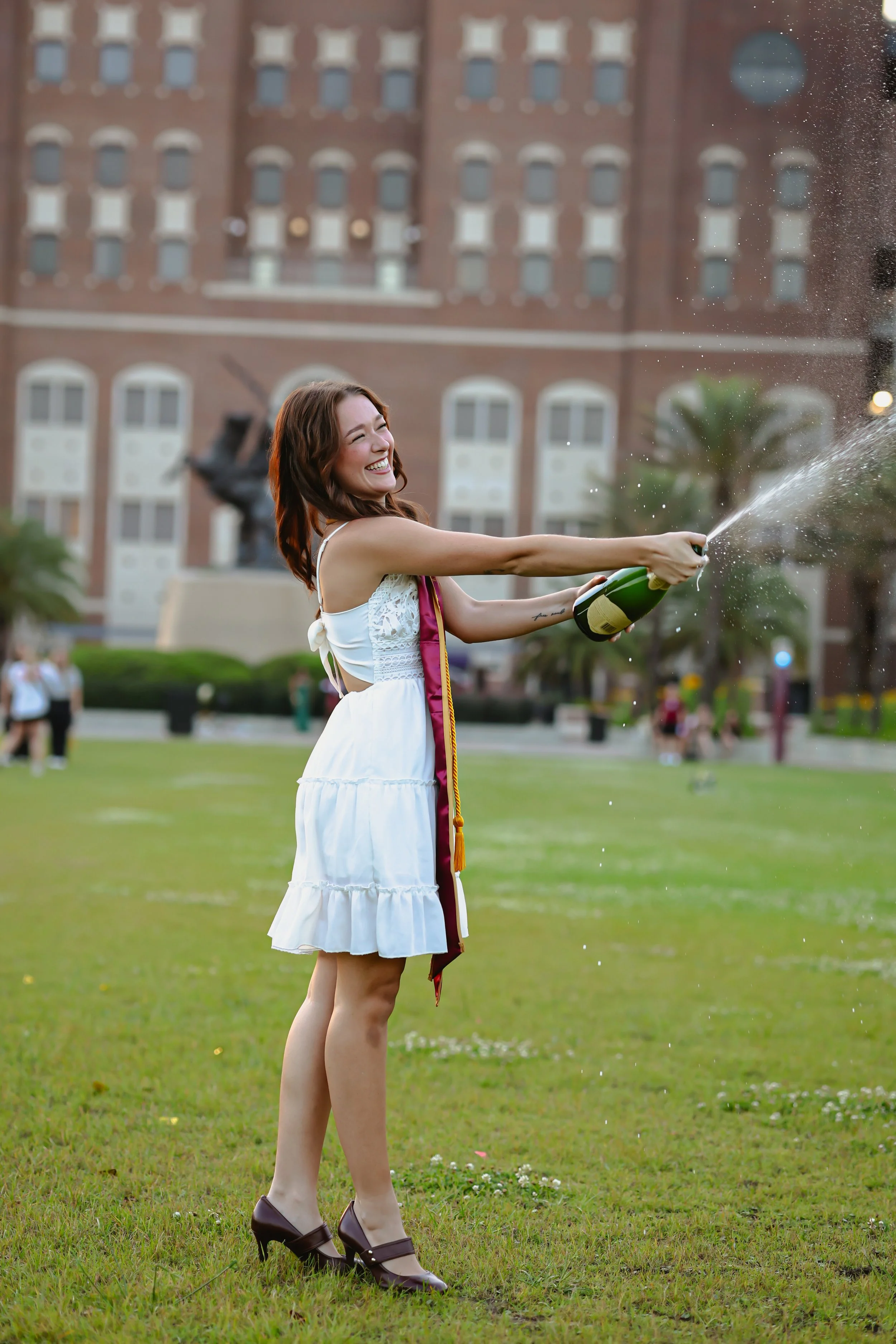 A young woman in a white dress celebrating her graduation outdoors, popping a champagne bottle with a big smile amidst a grassy area, with a large brick building in the background.