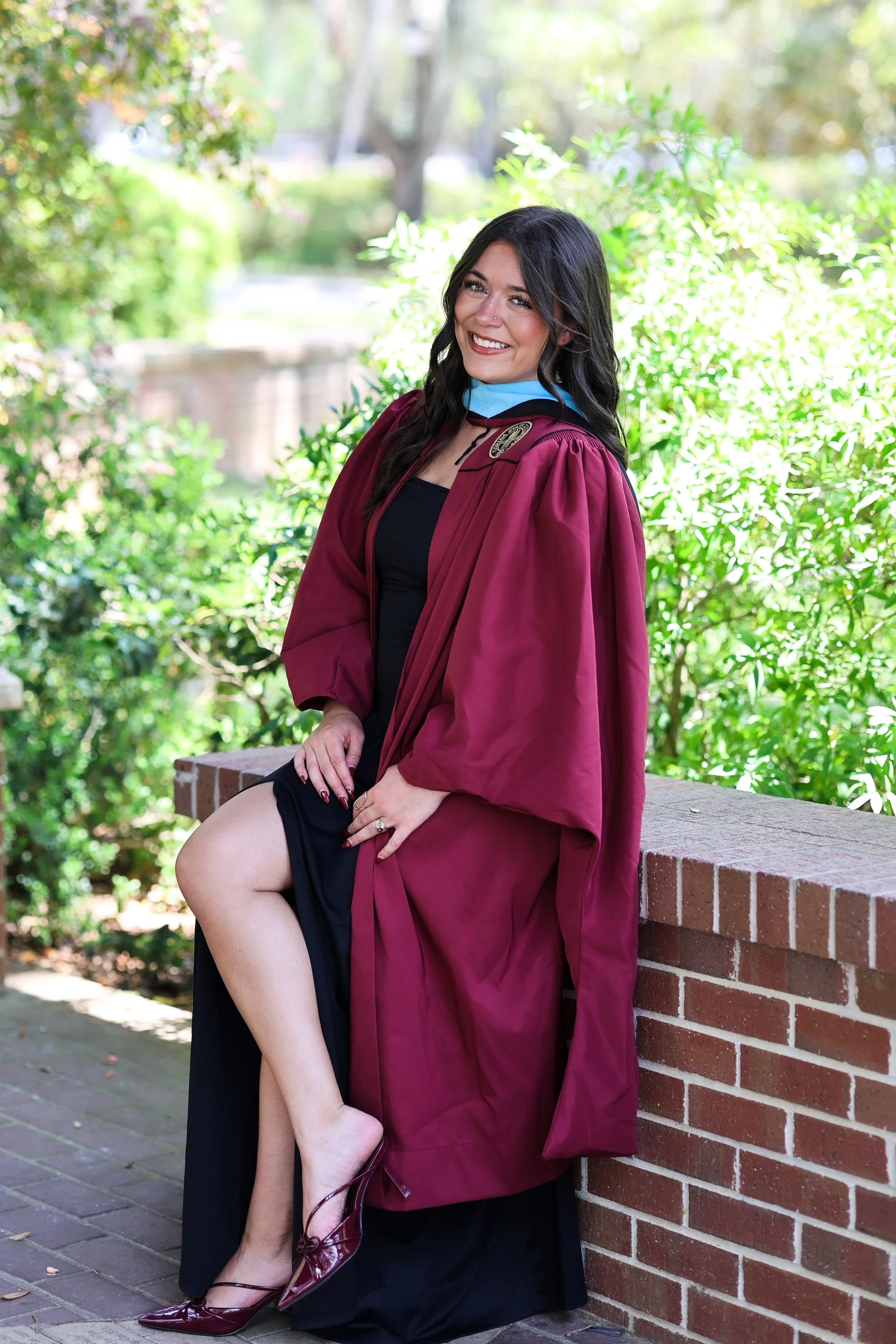 Woman in a graduation gown sitting on a brick ledge outdoors, smiling, with greenery in the background.