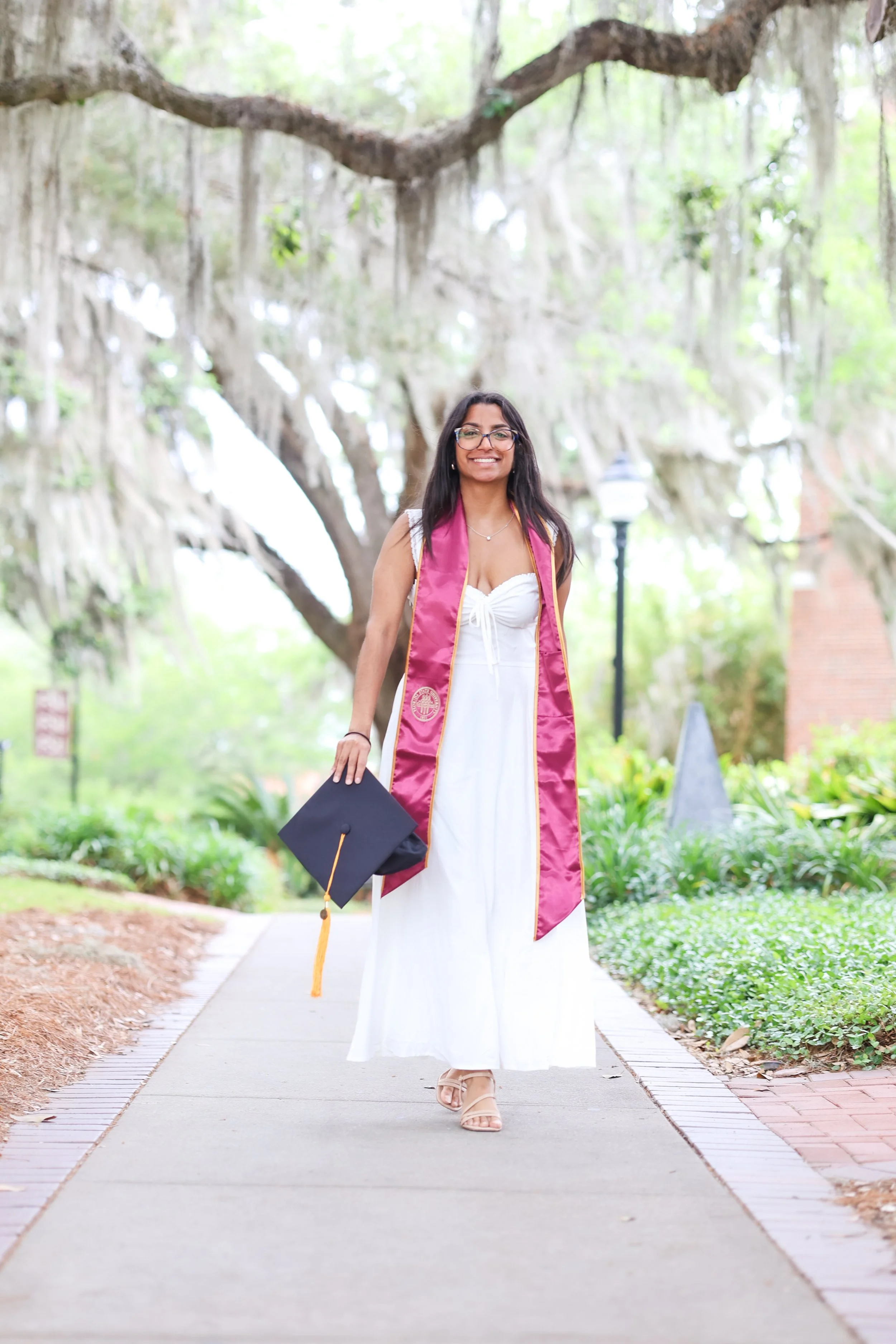 Young woman in a white dress and glasses, wearing a graduation stole and cap, walking on a pathway in a park with green trees and hanging moss.