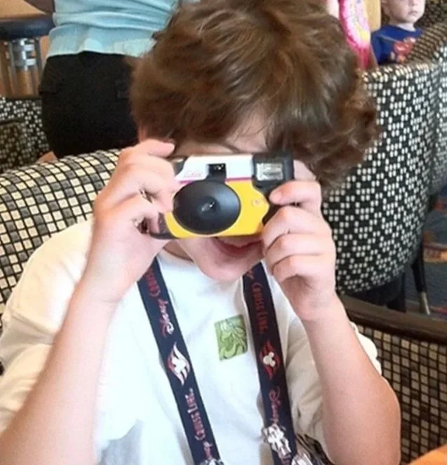 A young child with curly brown hair holding a plastic toy camera up to his face inside a room with patterned chairs and other children in the background.