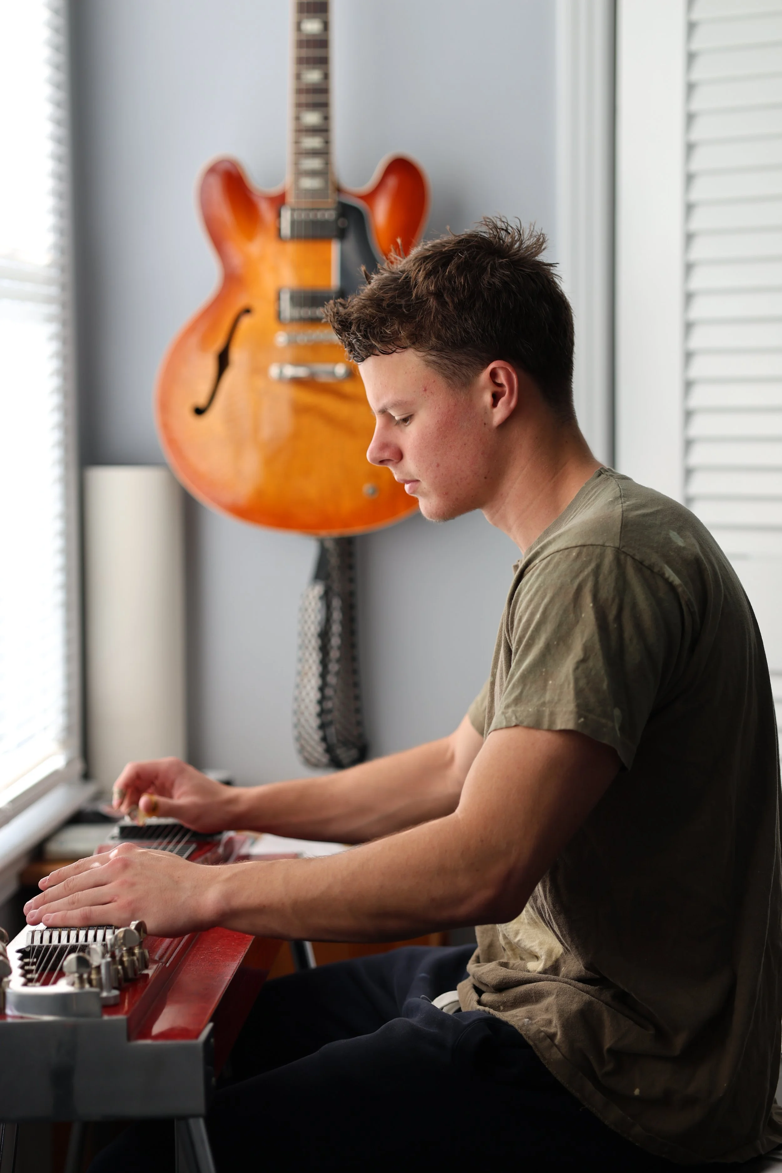 A young man with short, curly brown hair sitting at a red keyboard with his eyes closed, playing or practicing. Behind him hangs an orange semi-hollow electric guitar on a wall, next to a window with blinds.