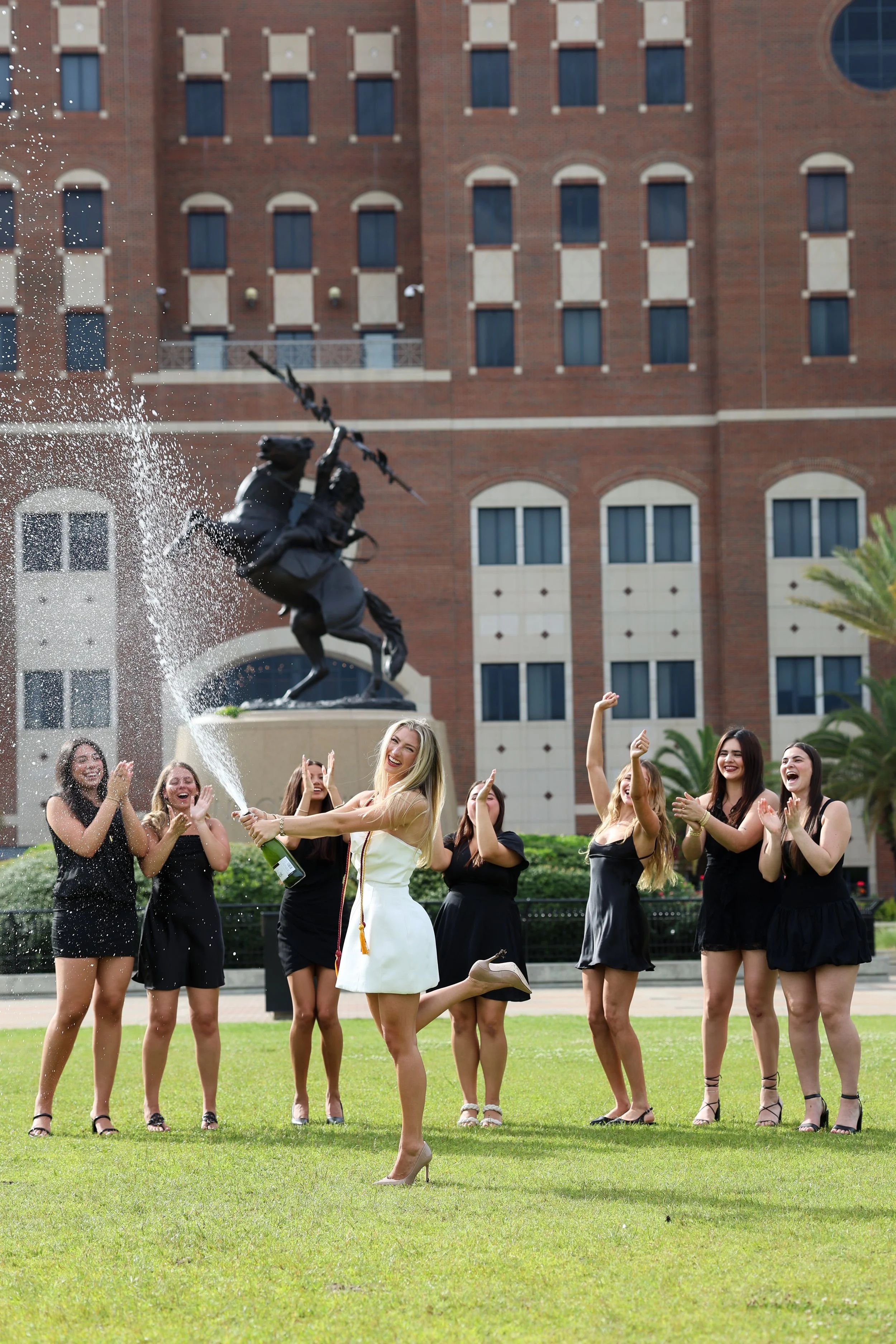 A celebration scene with a woman in a white dress popping champagne in front of a group of women in black dresses with their arms raised and smiling, on a green lawn with a fountain and a large brick building in the background.