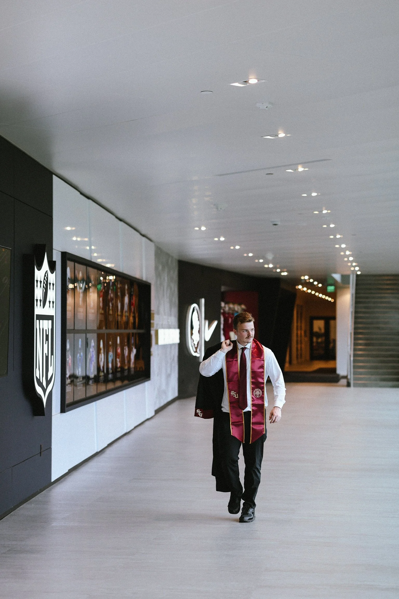 A young man dressed in formal attire, including a white shirt, red and gold stole, and black trousers, is walking in a modern indoor commercial space with a confident expression.