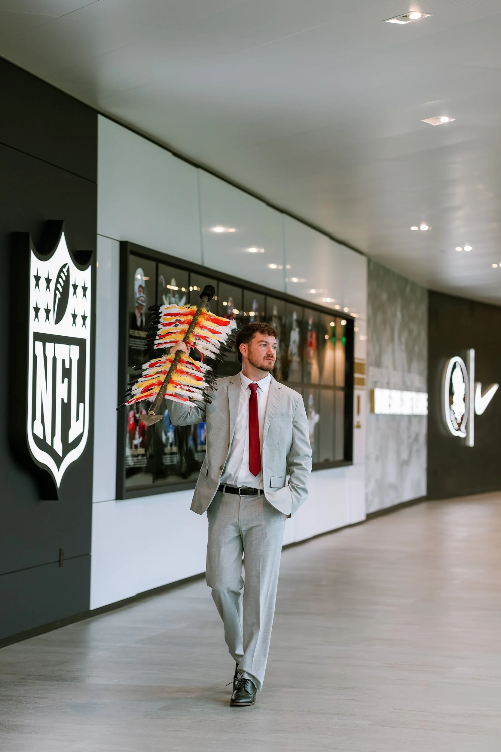 A man in a gray suit with a red tie is walking through a modern indoor space, carrying a colorful feathered pole. The background features a wall with a neon NHL logo and display cases.