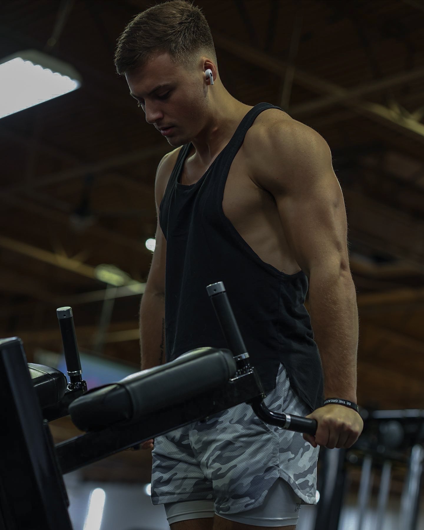 Young man in gym wearing black sleeveless shirt and camouflage shorts, holding a workout machine handle, focused on exercise.