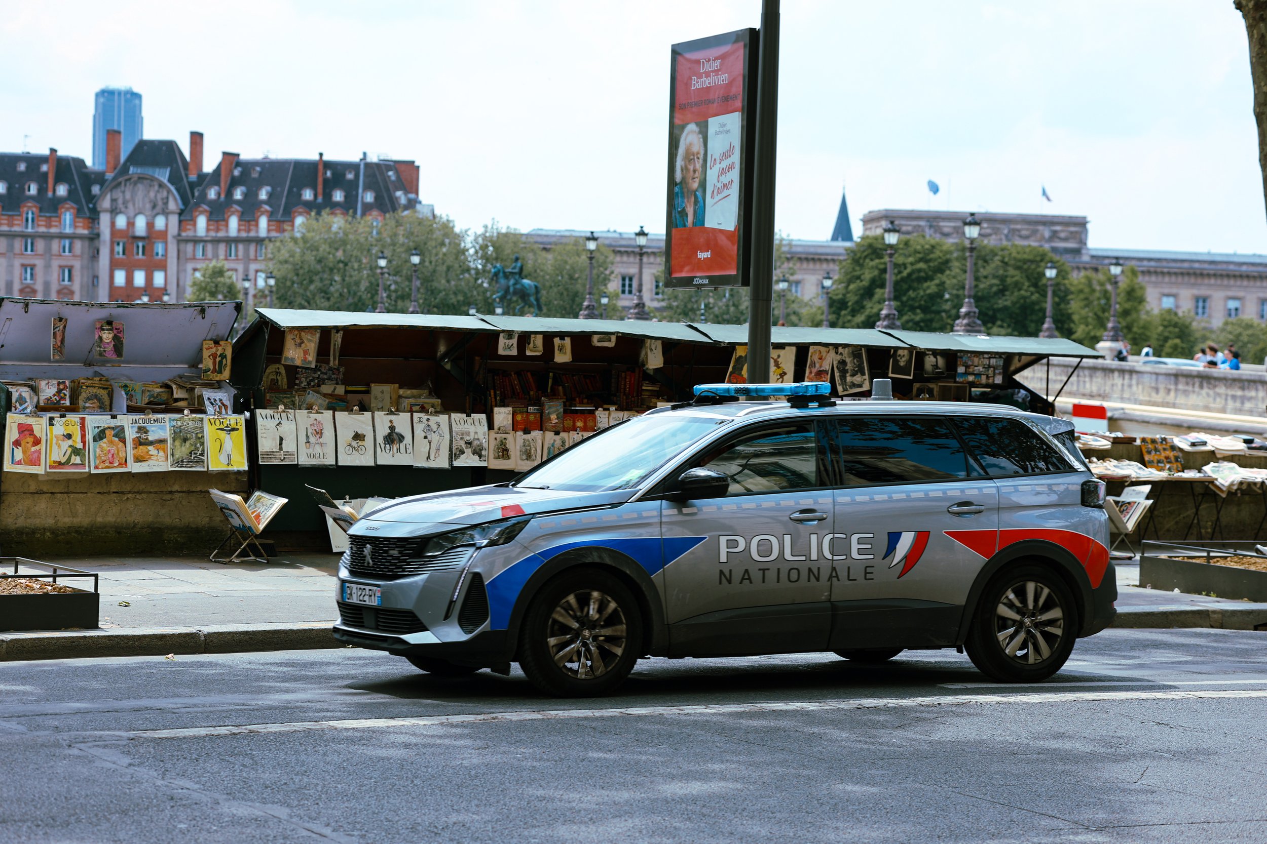 Paris police car parked near a street art and book stall with historic buildings and trees in the background.