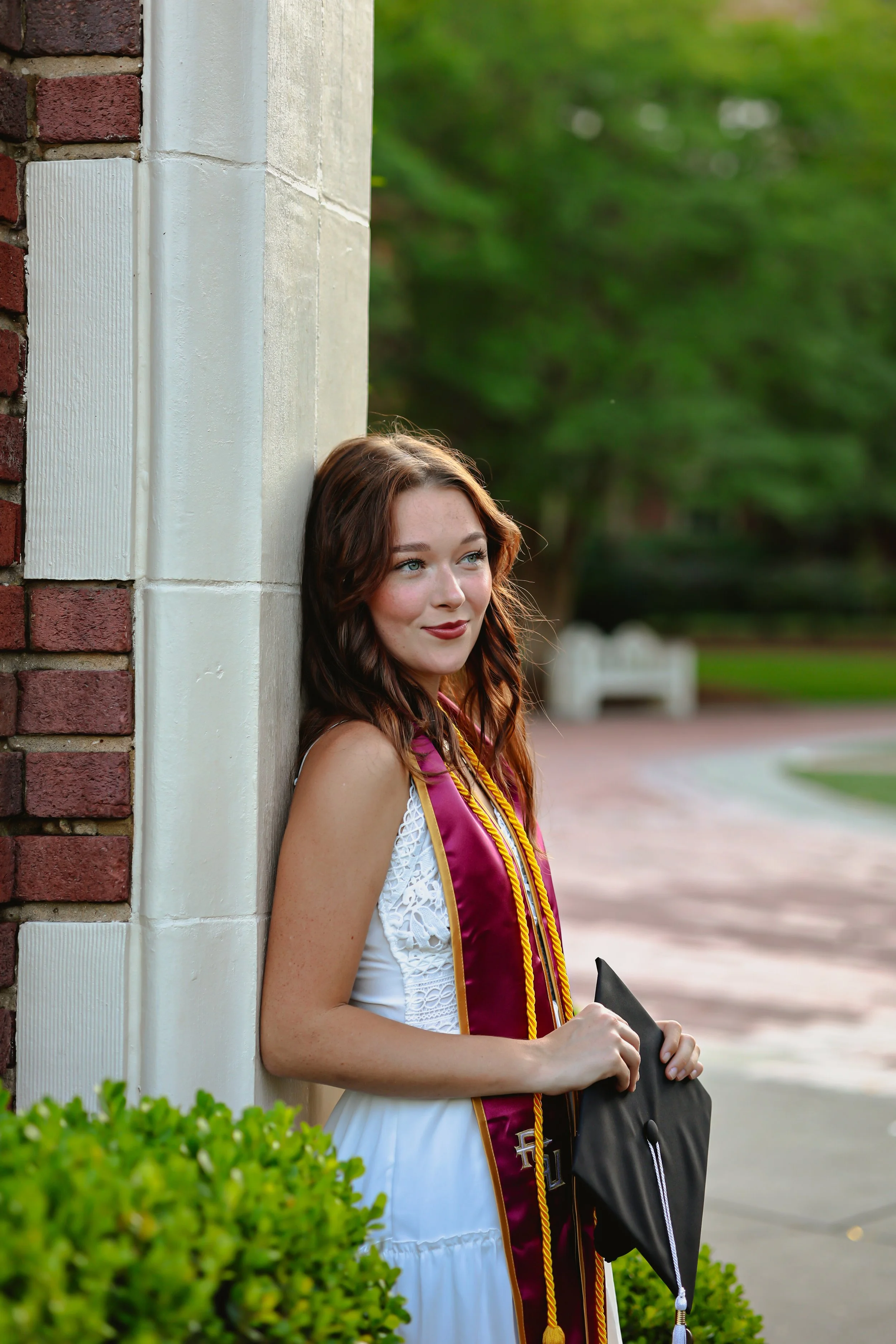 A young woman with brown hair and fair skin, wearing a white dress, stands outdoors beside a brick and stone wall, holding a graduation cap, with green trees and a pathway in the background.