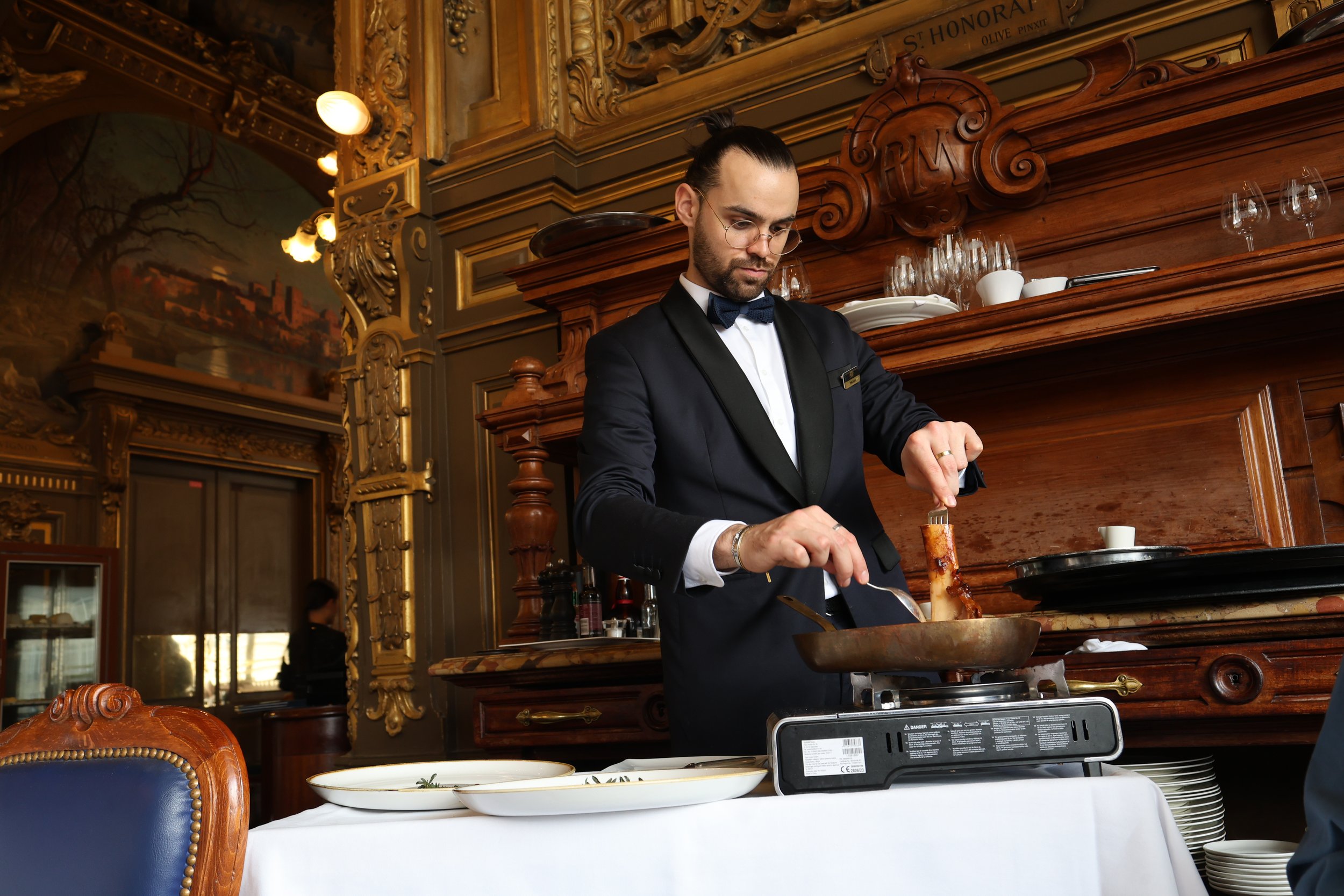 A male chef in a tuxedo garnished with a bow tie, cooking a dish in a luxurious, ornate restaurant with dark wood paneling and classical artwork.