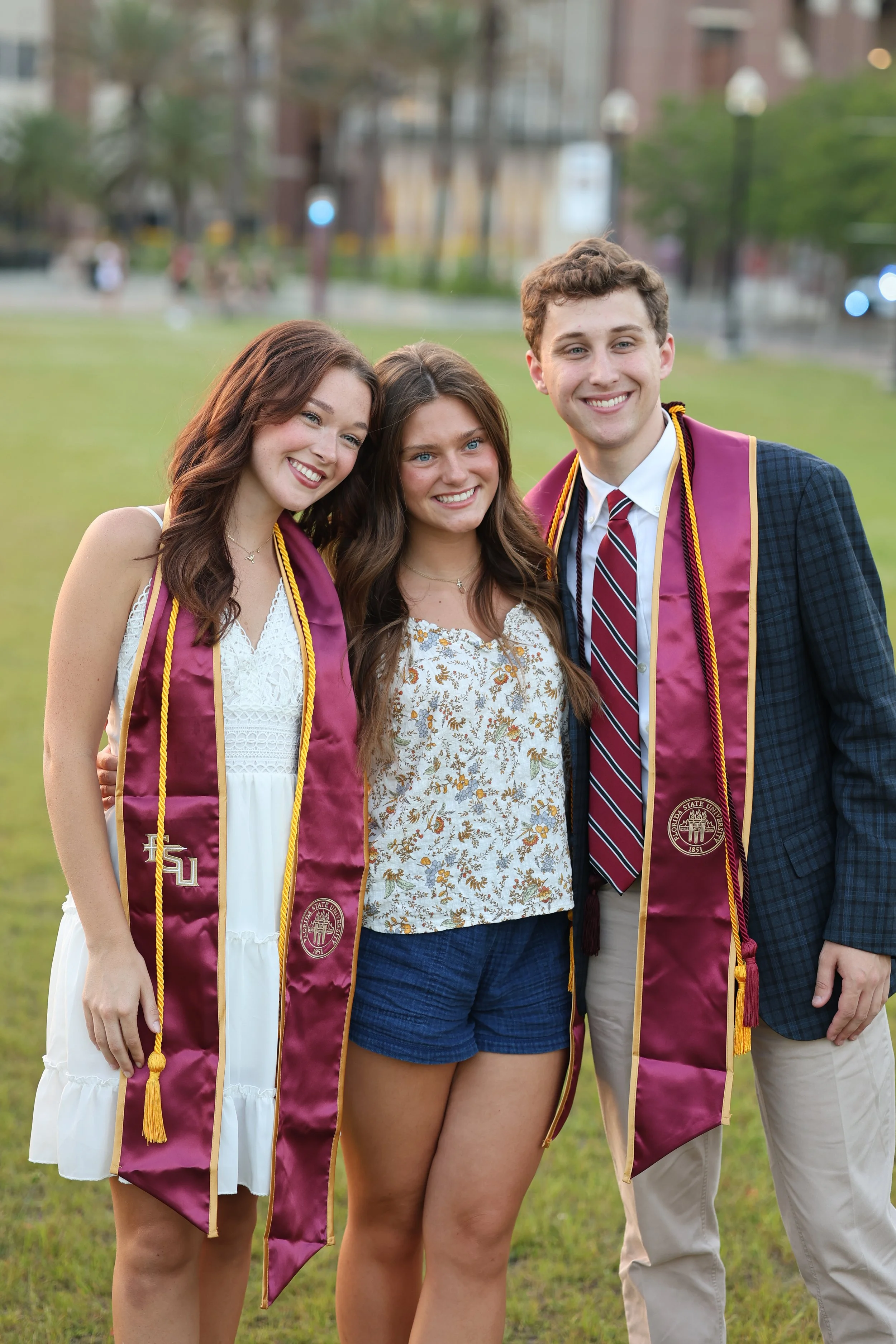 Three young adults, two women and one man, smiling and standing close together outdoors on a grassy field, with two of them wearing graduation stoles from Florida State University.