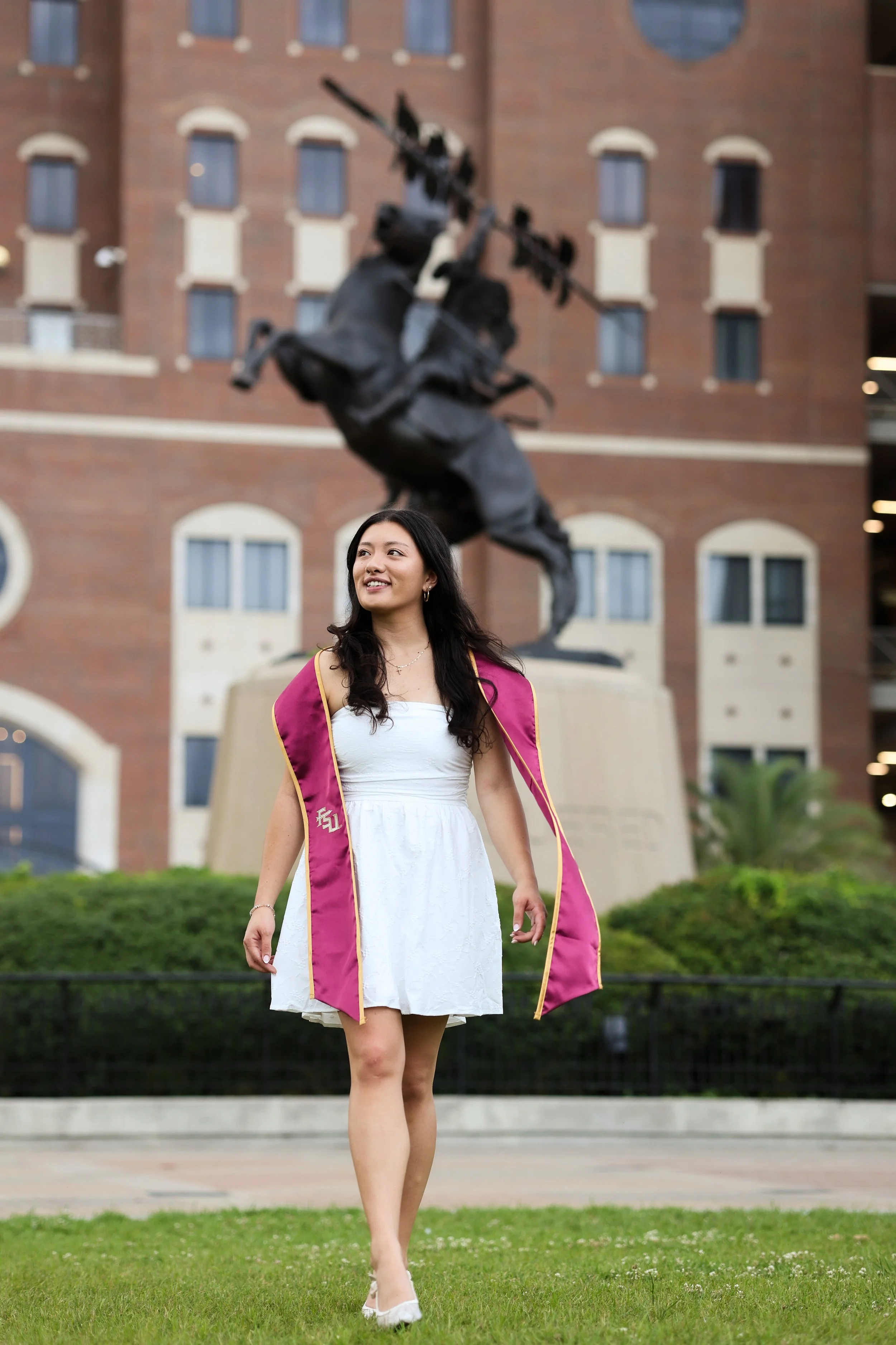 A young woman in a white dress and graduation cape walking on grass, with a large statue of a person on a horse in the background and a brick building behind the statue.
