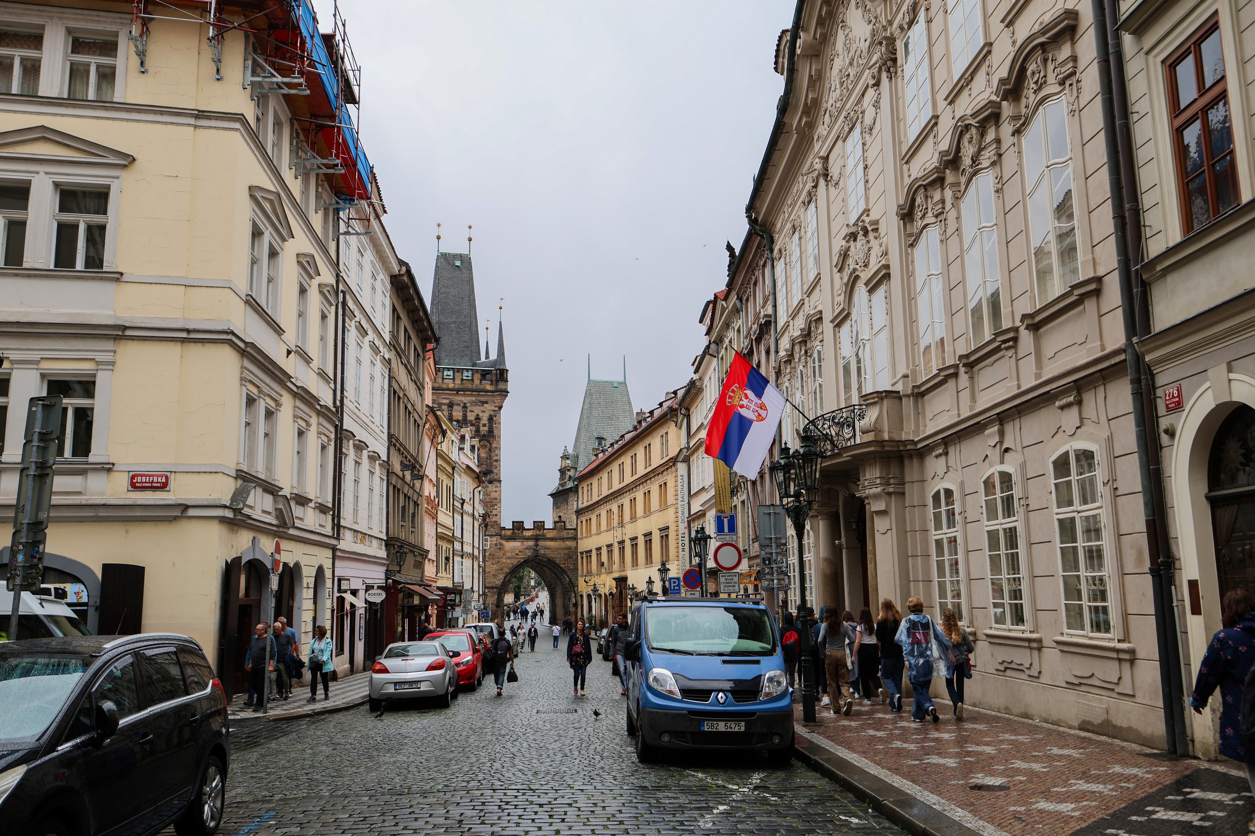 A historic European street with cobblestone pavement, lined with ornate old buildings, some under renovation, and a Serbian flag hanging outside one building. Pedestrians walk along the sidewalks, and a stone archway with a tower is visible in the background.