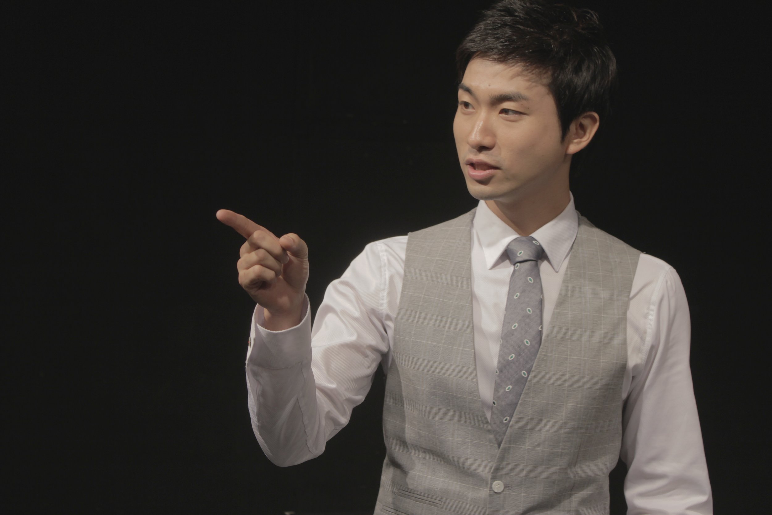 A young man in formal attire, white shirt, grey vest, and polka dot tie, pointing to his left against a black background.