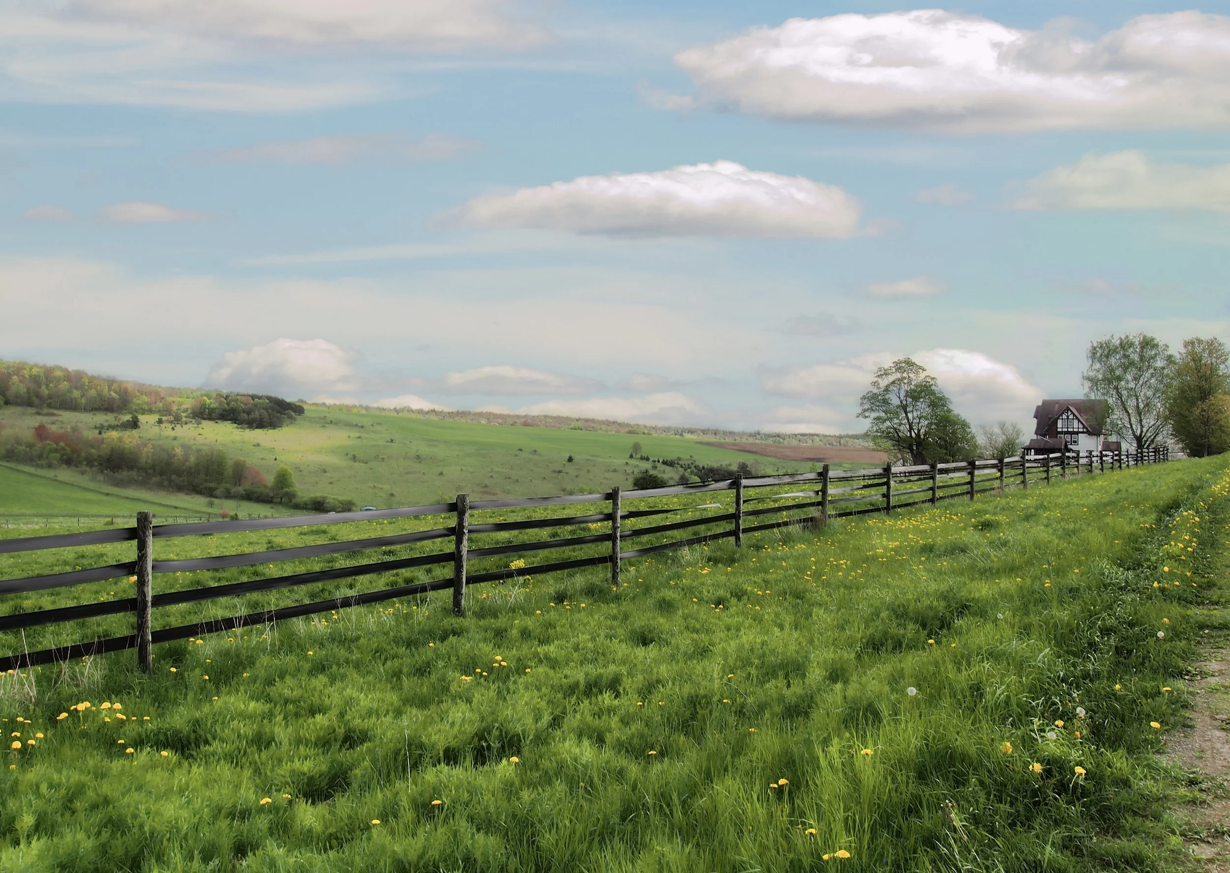 A scenic countryside landscape with a green grassy field, a wooden fence, rolling hills in the distance, a house among trees, and a partly cloudy sky.