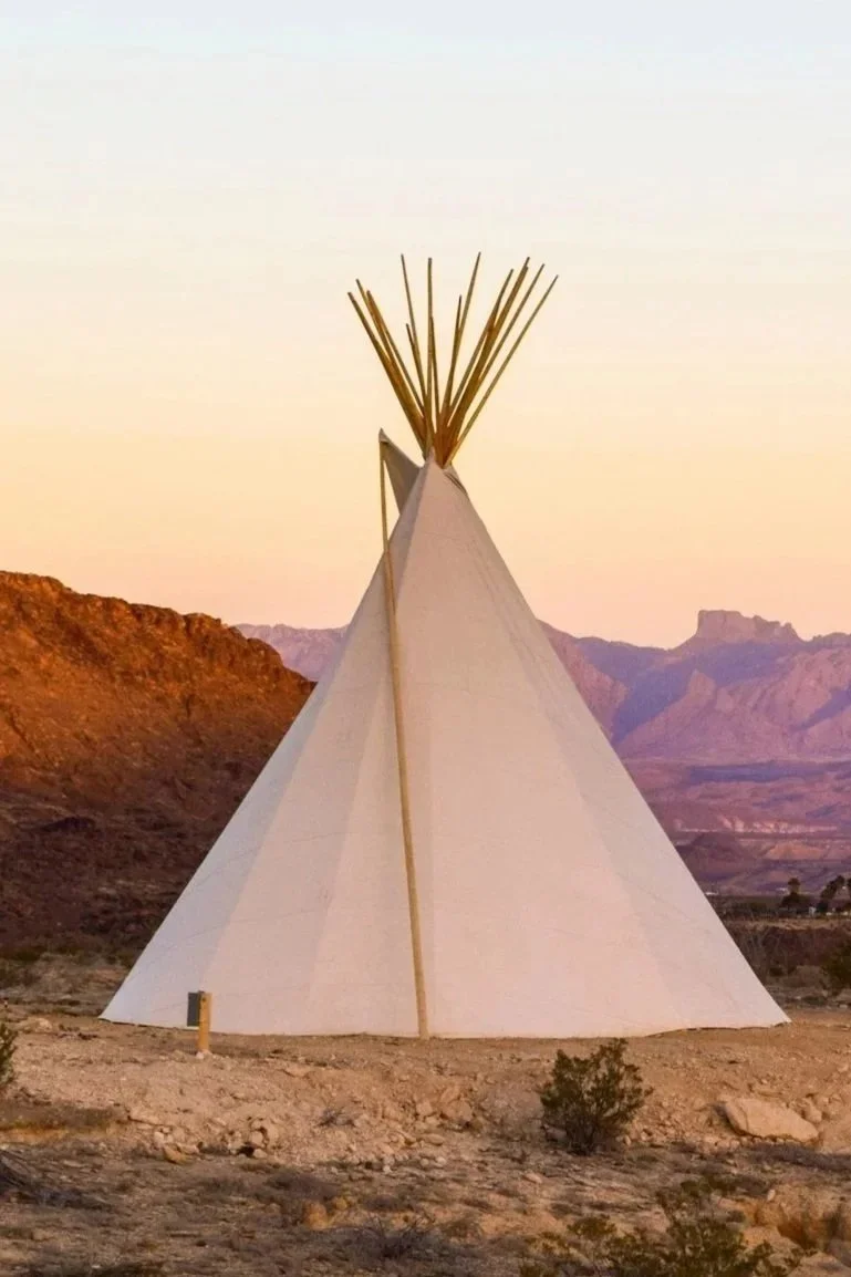 A teepee set up in a desert landscape with mountains in the background during sunset.
