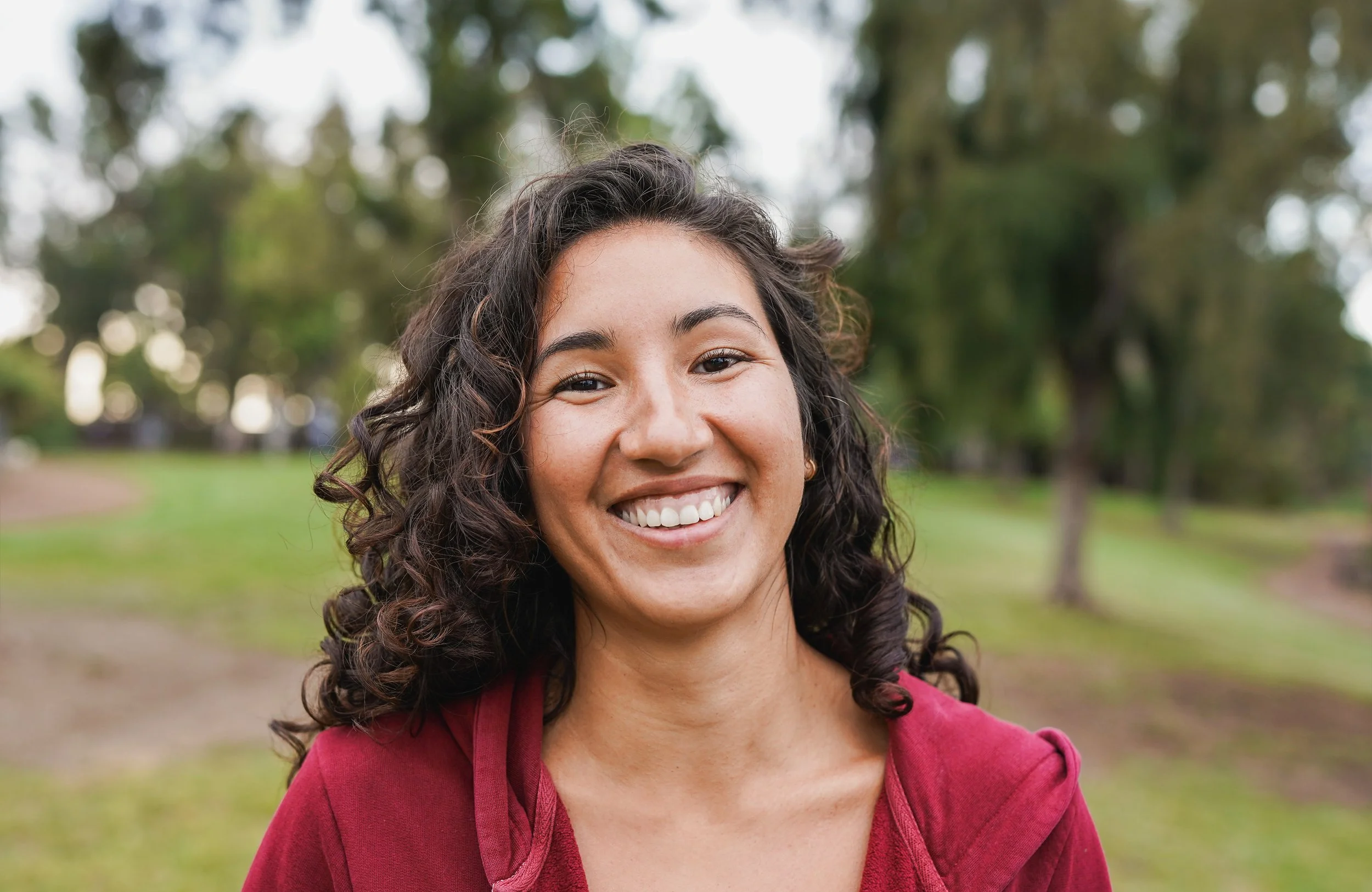 Close-up of a smiling young woman with curly dark hair outdoors.”