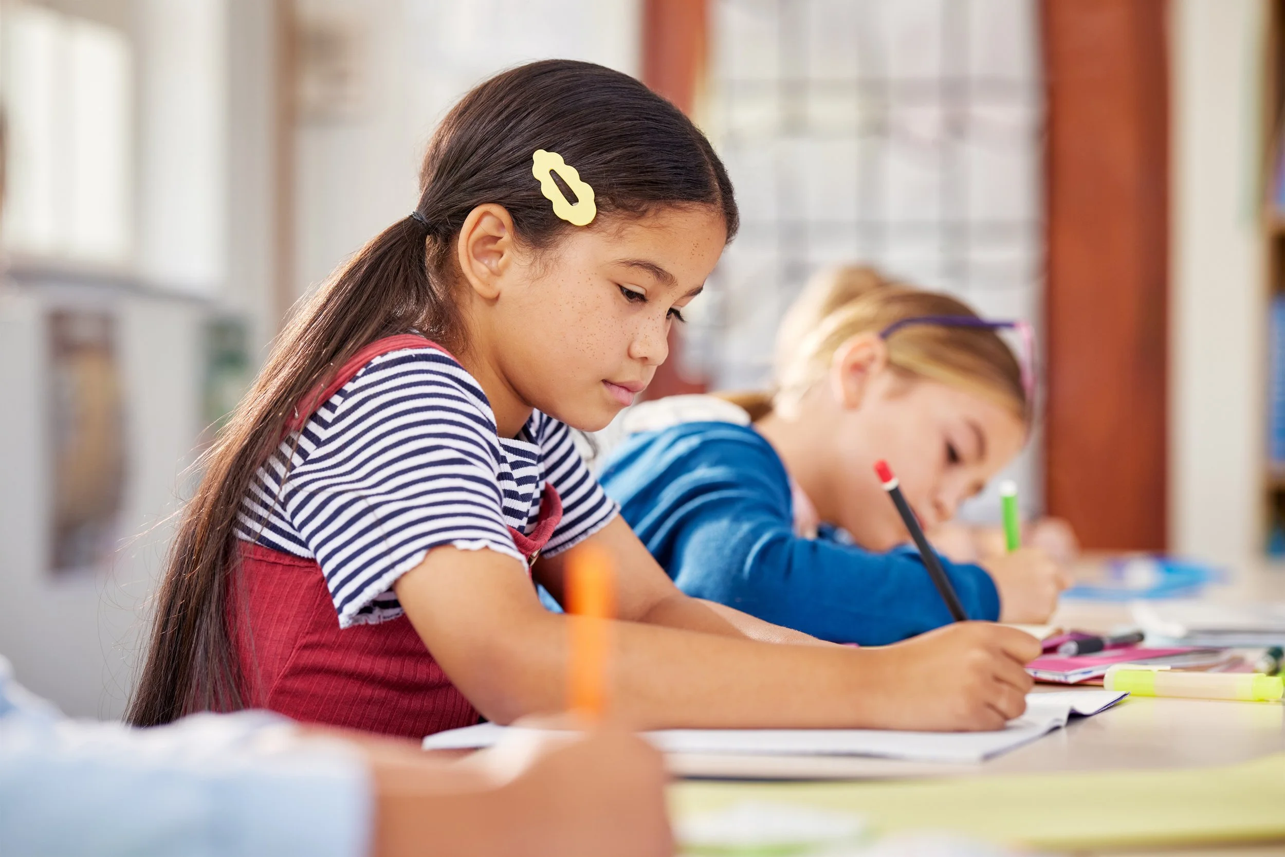 Two elementary school girls writing in notebooks at a classroom desk.