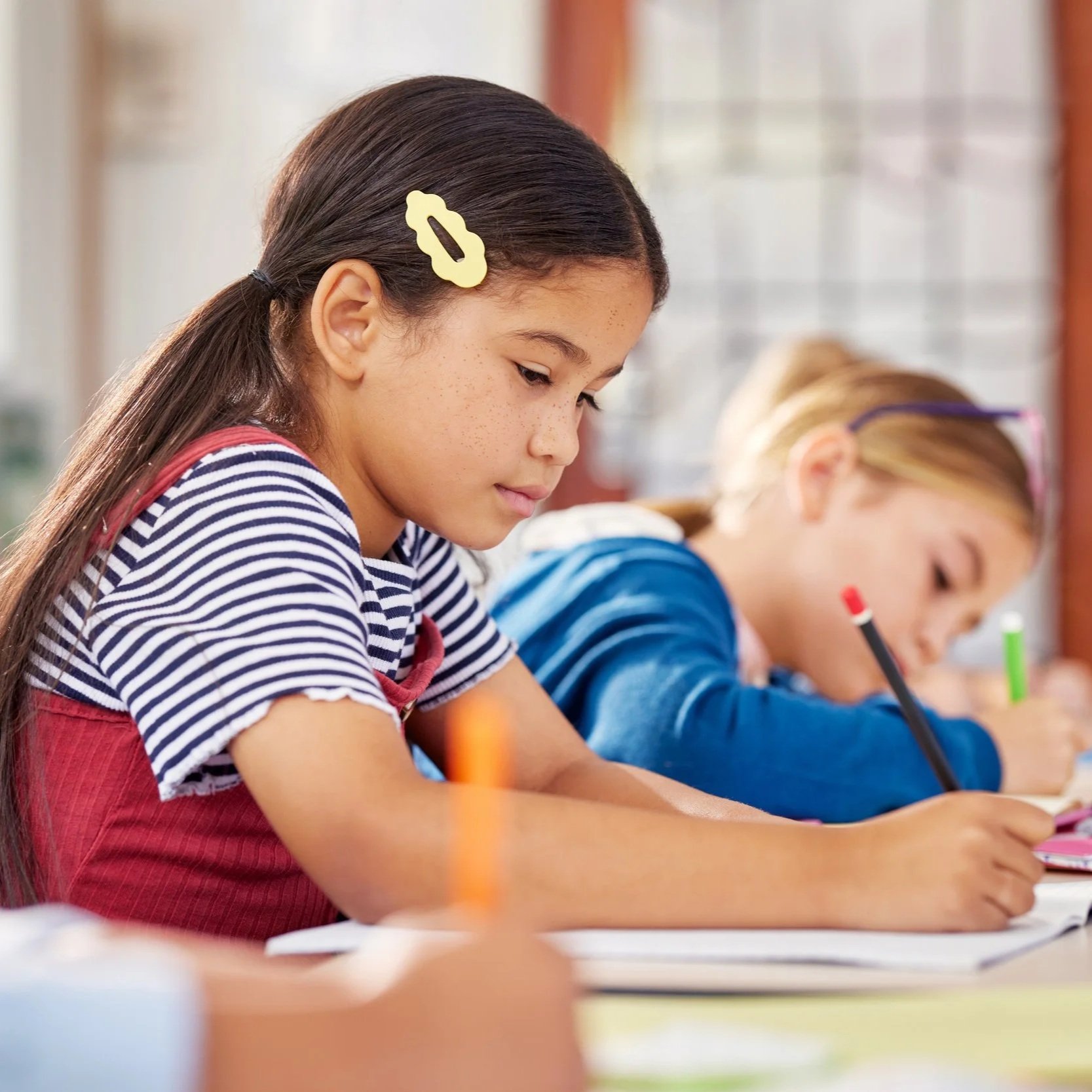Two young girls sitting at a desk, writing in notebooks, in a classroom setting.