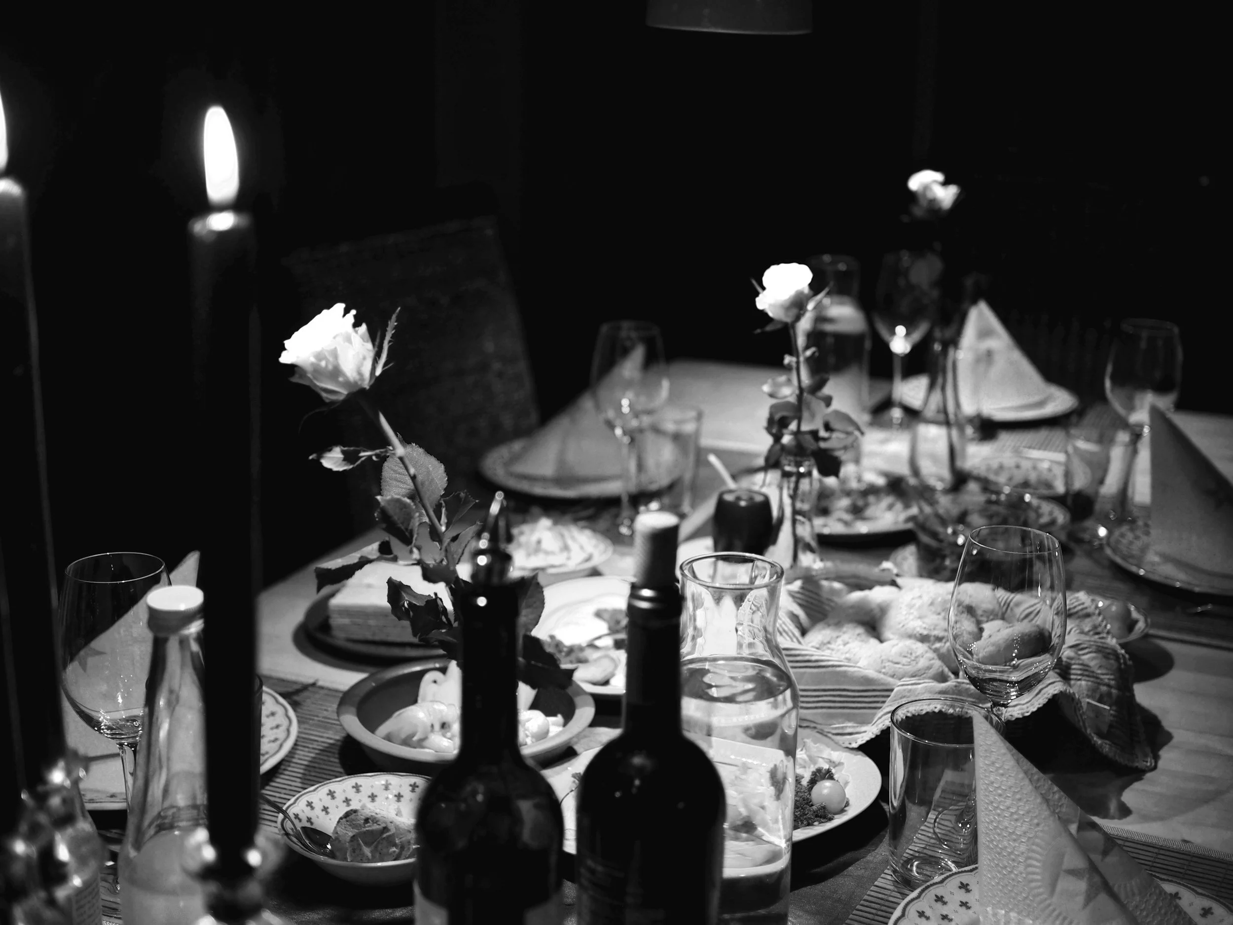 A dinner table setting with plates, glasses, and bottles. There are flowers in vases and bread, salad, and other dishes on the table. Candlelight illuminates the scene in black and white.