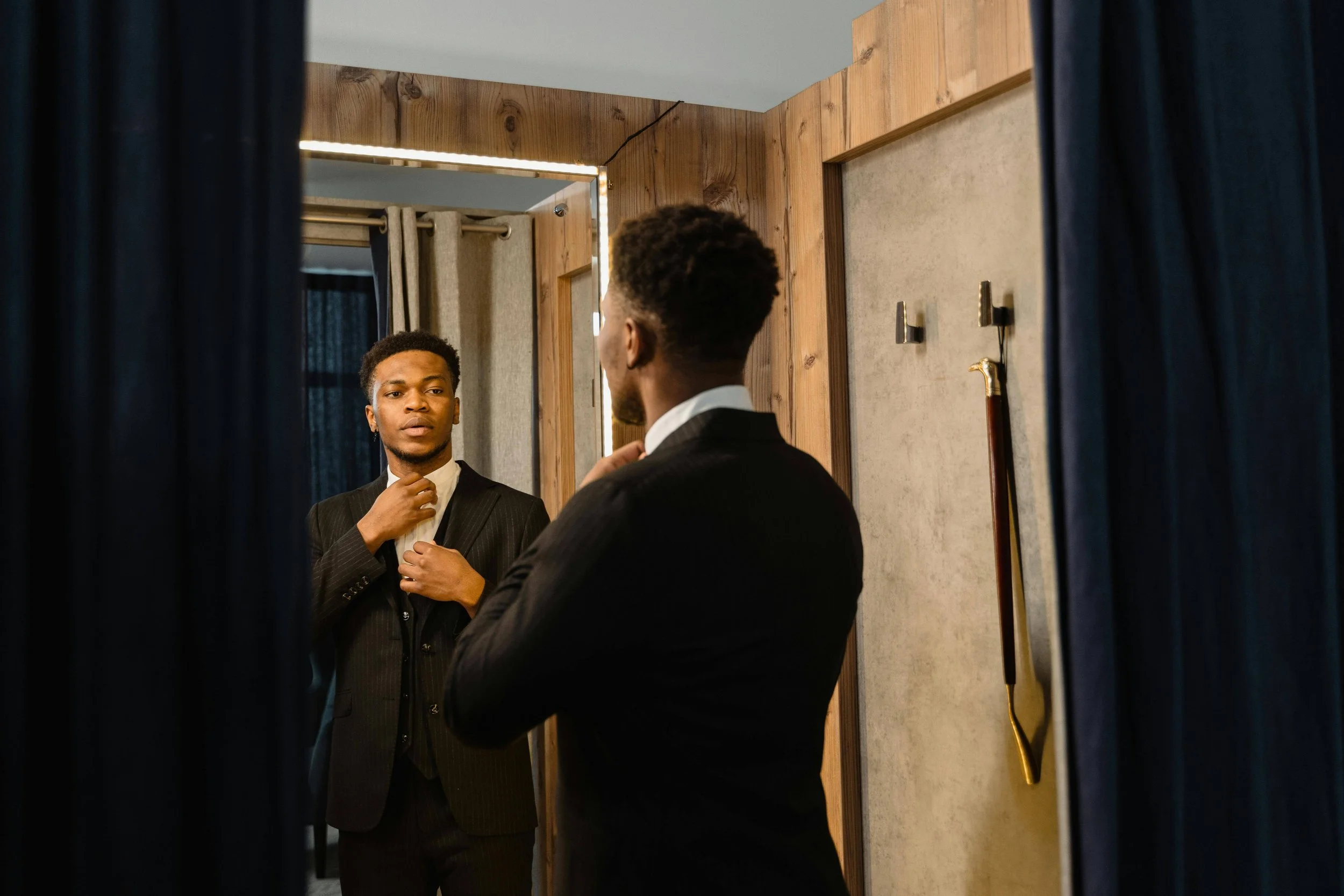 A man in a black suit looking at himself in a mirror, adjusting his tie, in a fitting room with wooden walls and a belt hanging on the wall.