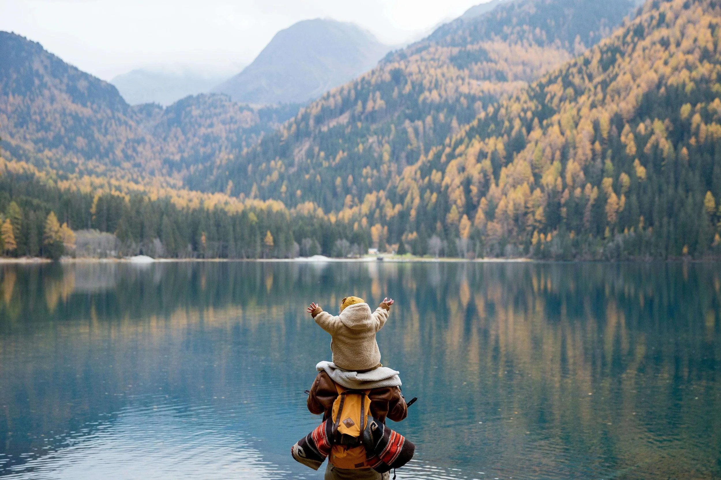 Child in a tan jacket and hat sitting on an adult's shoulders with arms raised, overlooking a lake surrounded by autumn-colored trees and mountains in the distance.