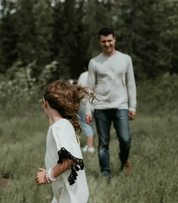 A girl with long hair running in a grassy field while two people, a man and a girl, walk behind her.