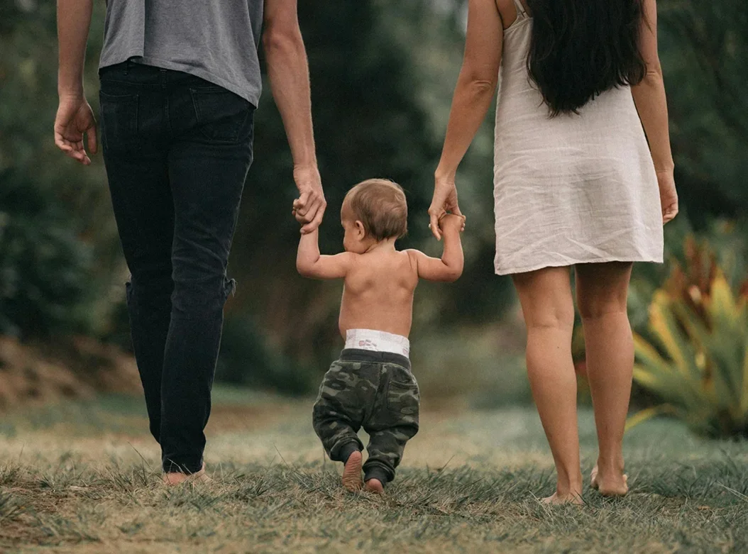 A young boy holding hands with two adults, one on each side, walking barefoot outdoors in a grassy area.