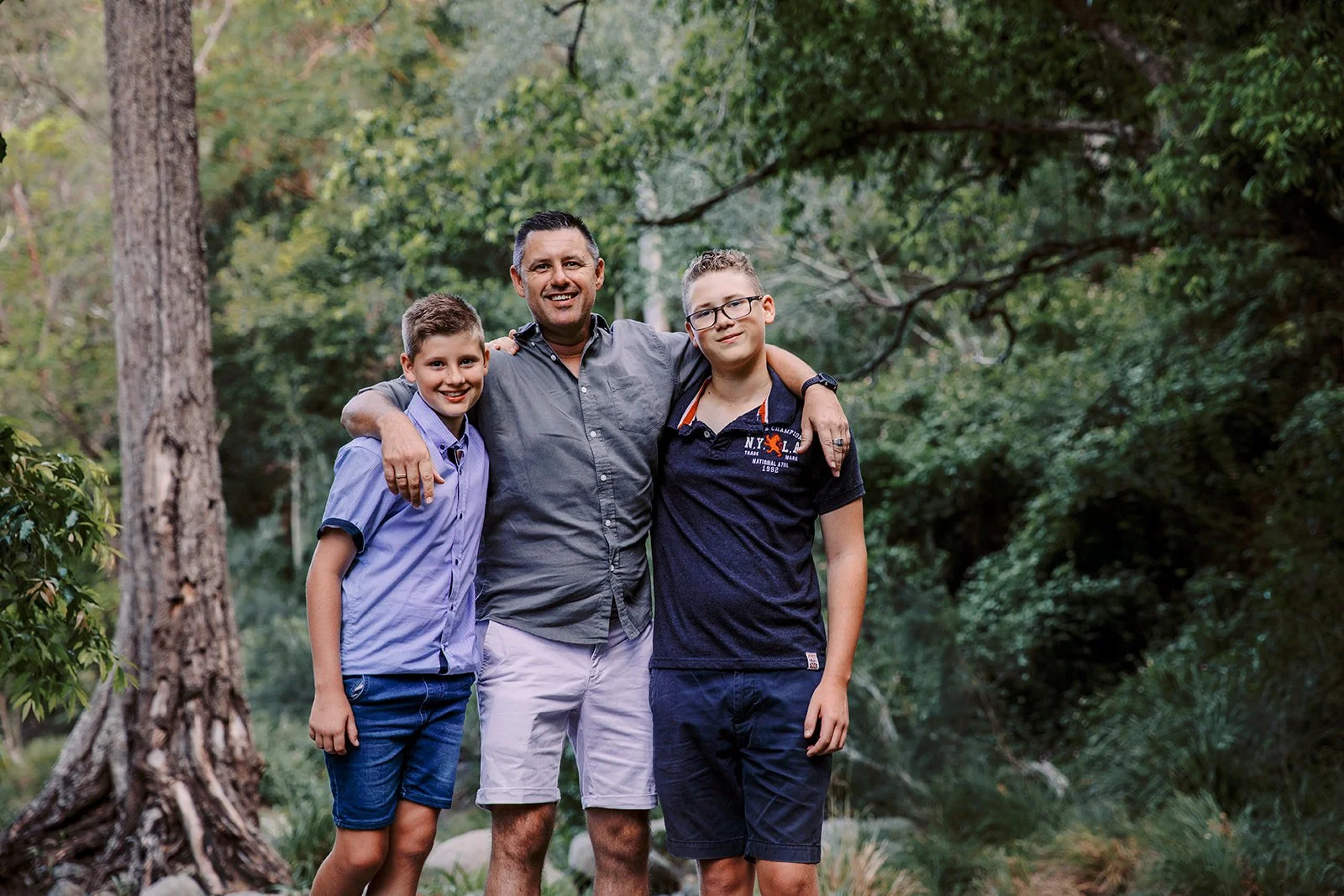 A man with two boys standing outdoors in a green wooded area, smiling at the camera.