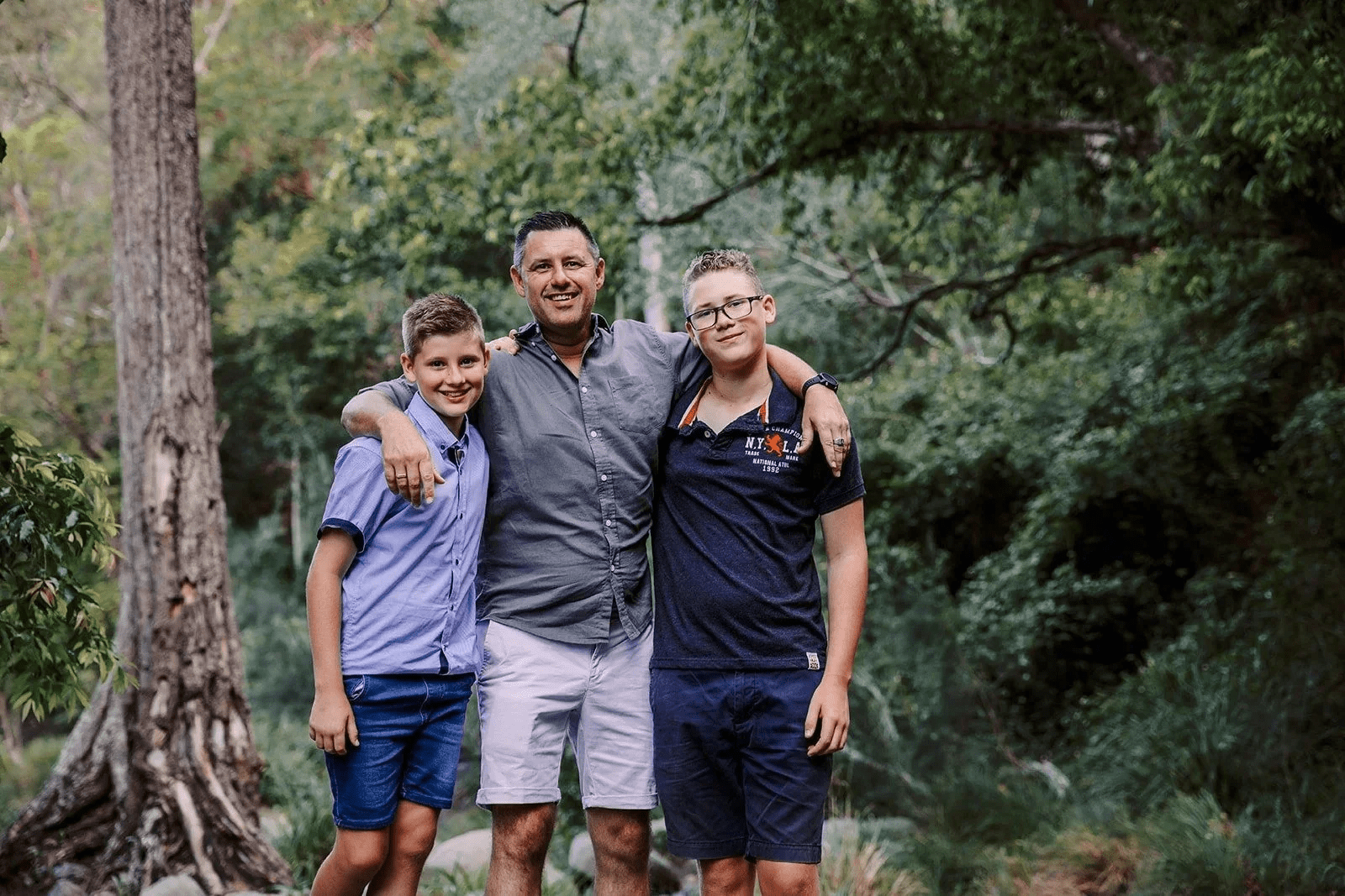 A man and two boys standing outdoors in a wooded area, smiling and with their arms around each other.