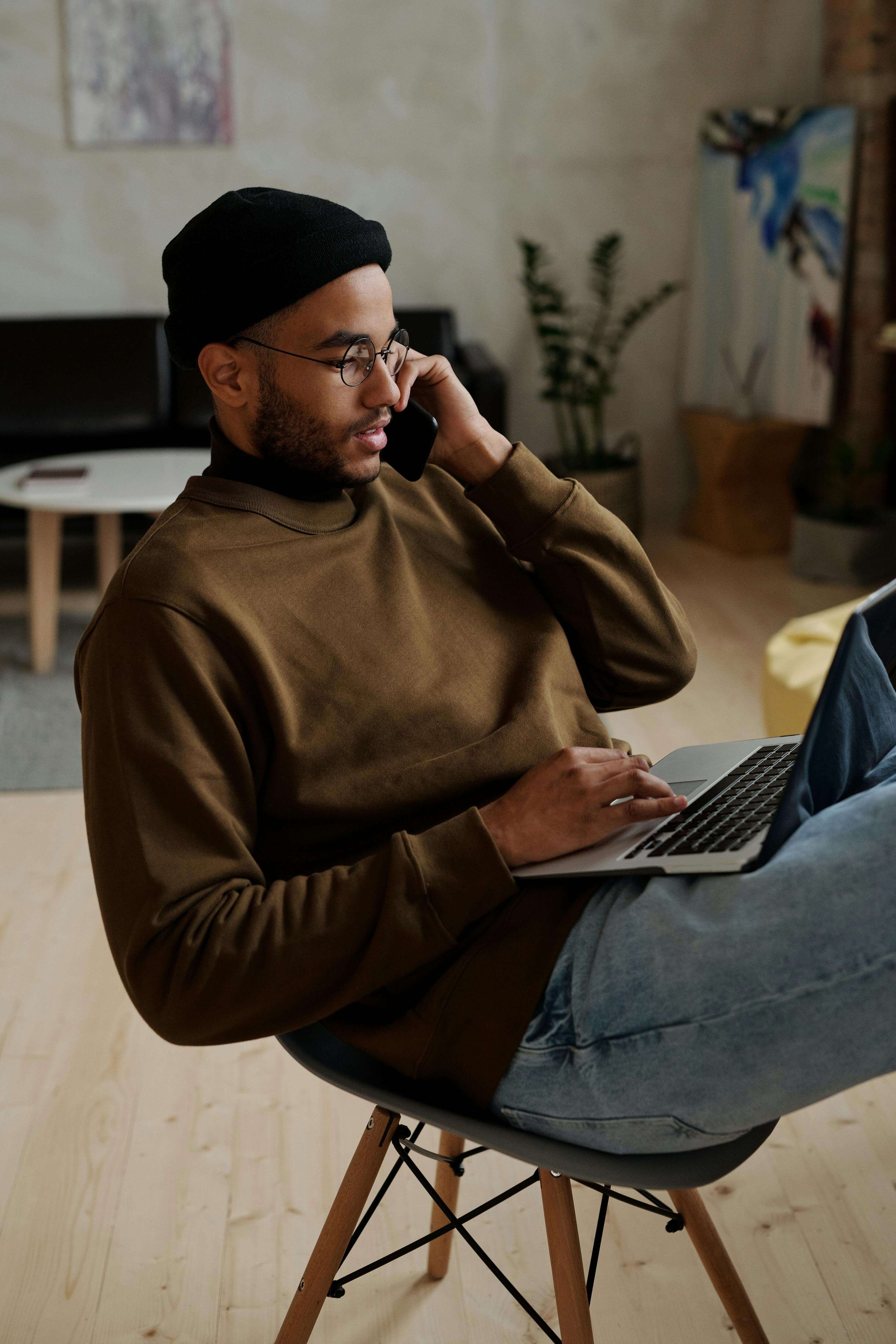 A man with glasses and a black beanie sitting on a modern chair, talking on a cellphone and using a laptop in a cozy room with art and plants.