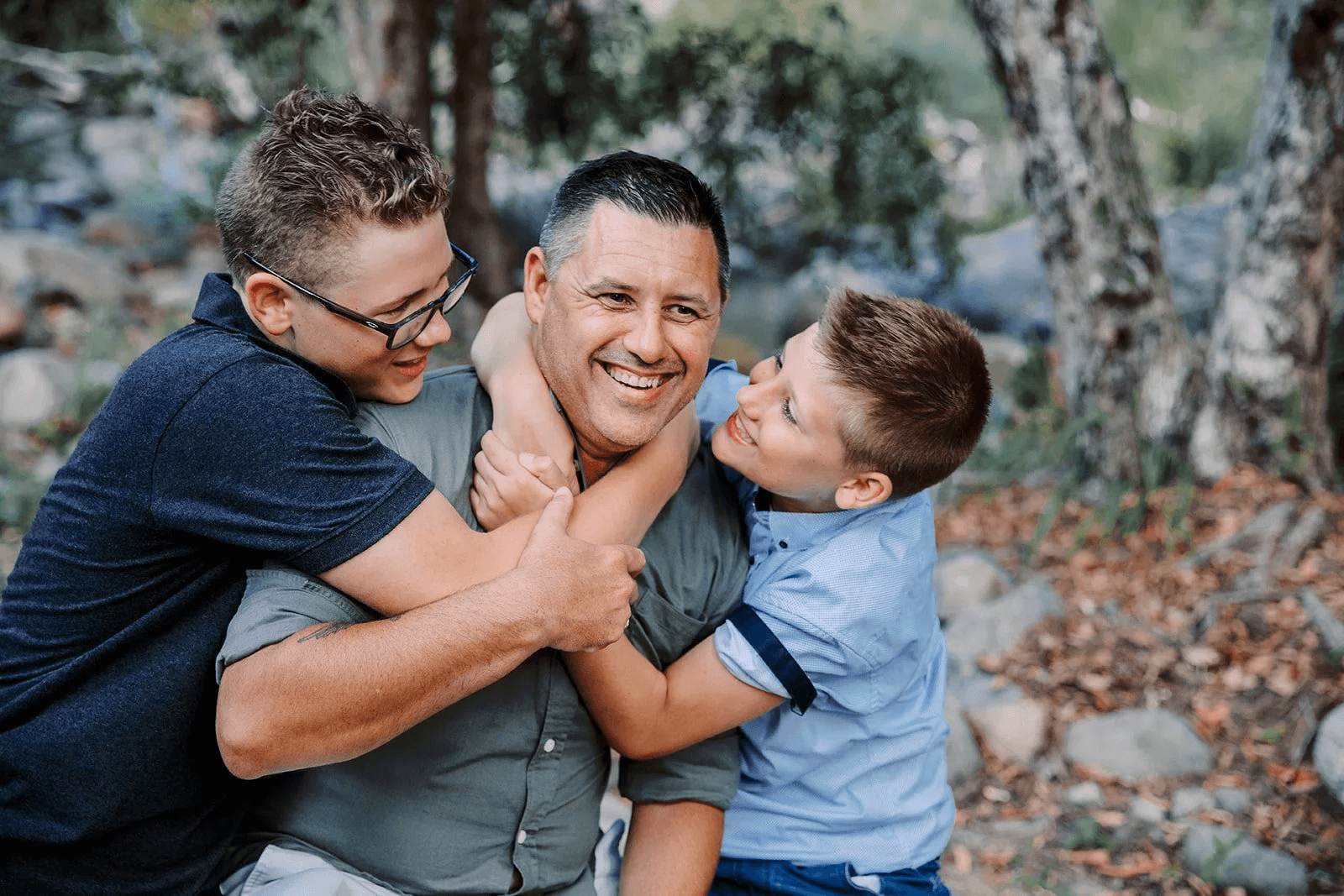 A joyful man in a gray shirt is playing with two young boys outdoors among trees and rocks. One boy, in glasses and a navy shirt, is hugging the man, while the other, in a light blue shirt, is smiling and holding onto the man's neck. All are smiling and enjoying the moment.