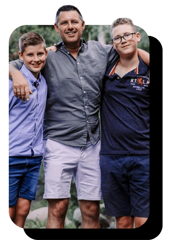 Three males standing outdoors, smiling, with arms around each other's shoulders. The man in the middle has short dark hair, a dark gray shirt, and light-colored shorts. The boy on the left has short brown hair and is wearing a purple button-up shirt with blue shorts. The boy on the right has light hair, glasses, a navy blue polo shirt, and navy shorts.